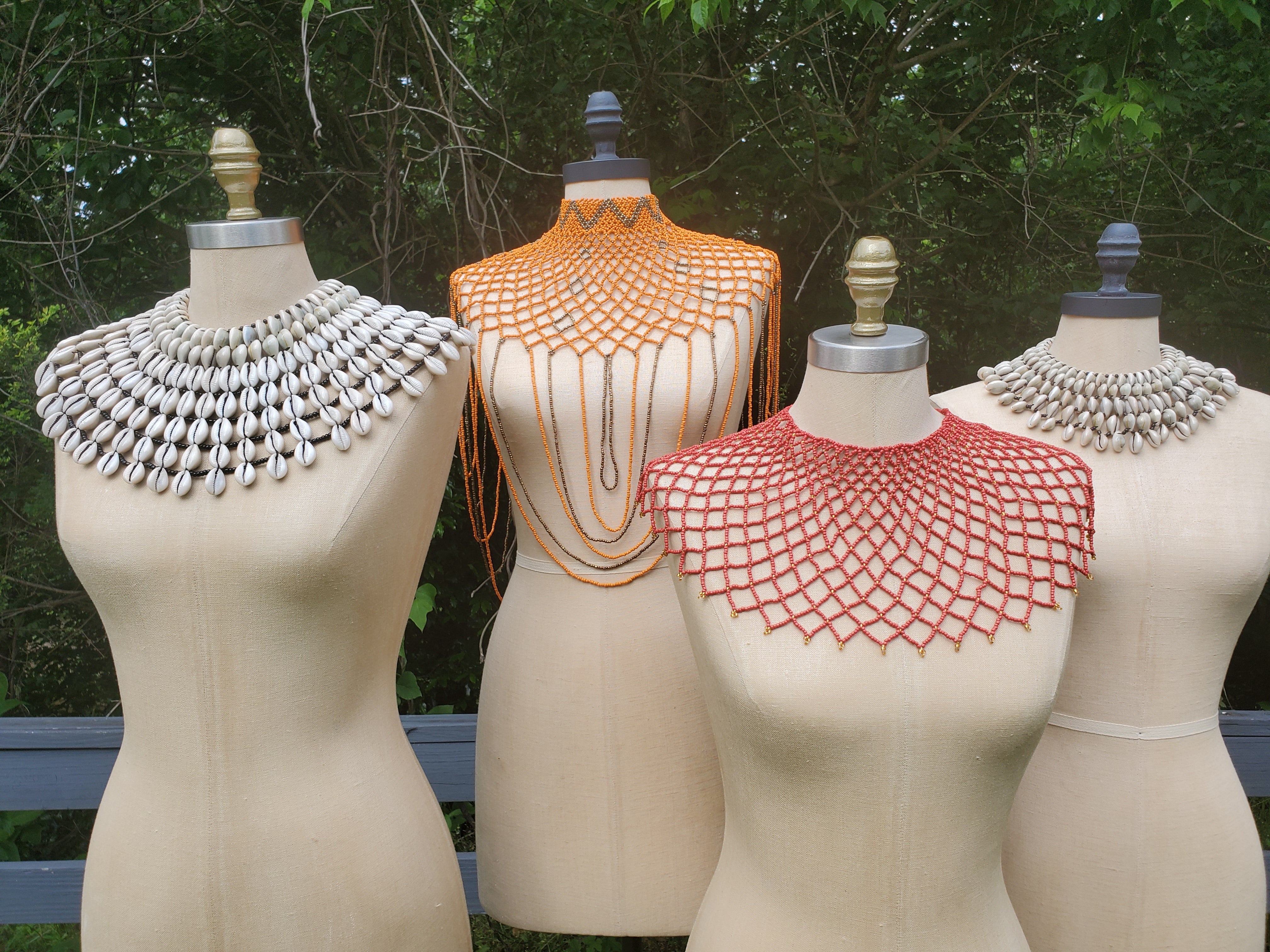 Four Shop Kamawe necklaces. From left to right our Large Reversible Cowrie Shell Necklace, an Orange and Gold Beaded Body Necklace, a Burnt Orange and Gold Beaded Collar Necklace and our Small Cowrie Shell Necklace.