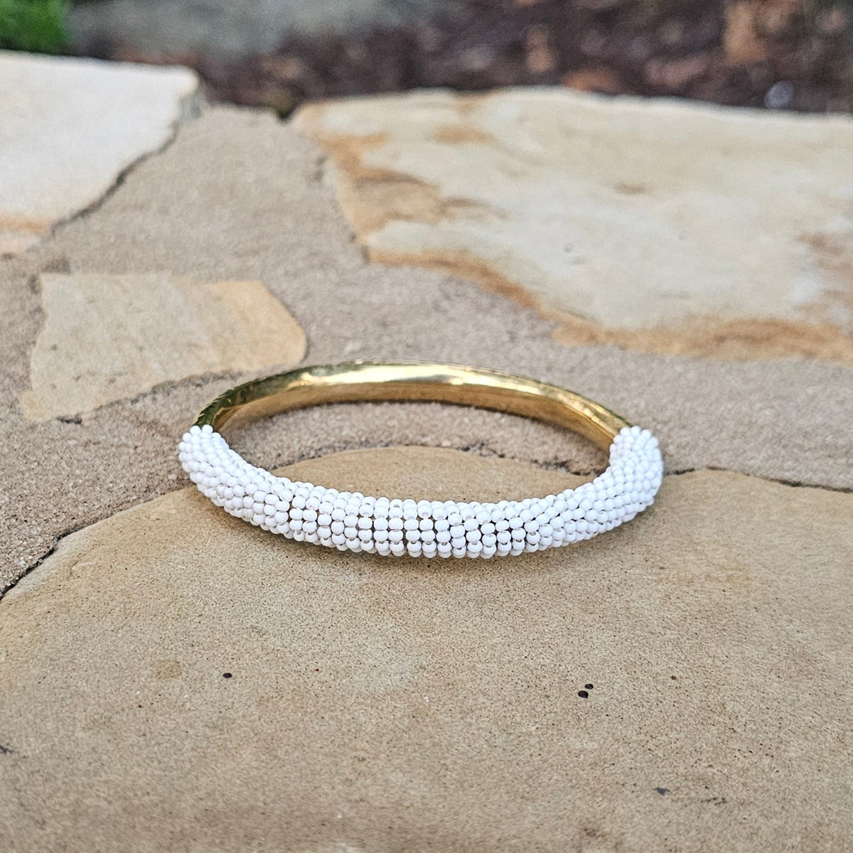 A gold bangle bracelet with a textured white beaded wrap, resting on a stone wall with green foliage in the background.