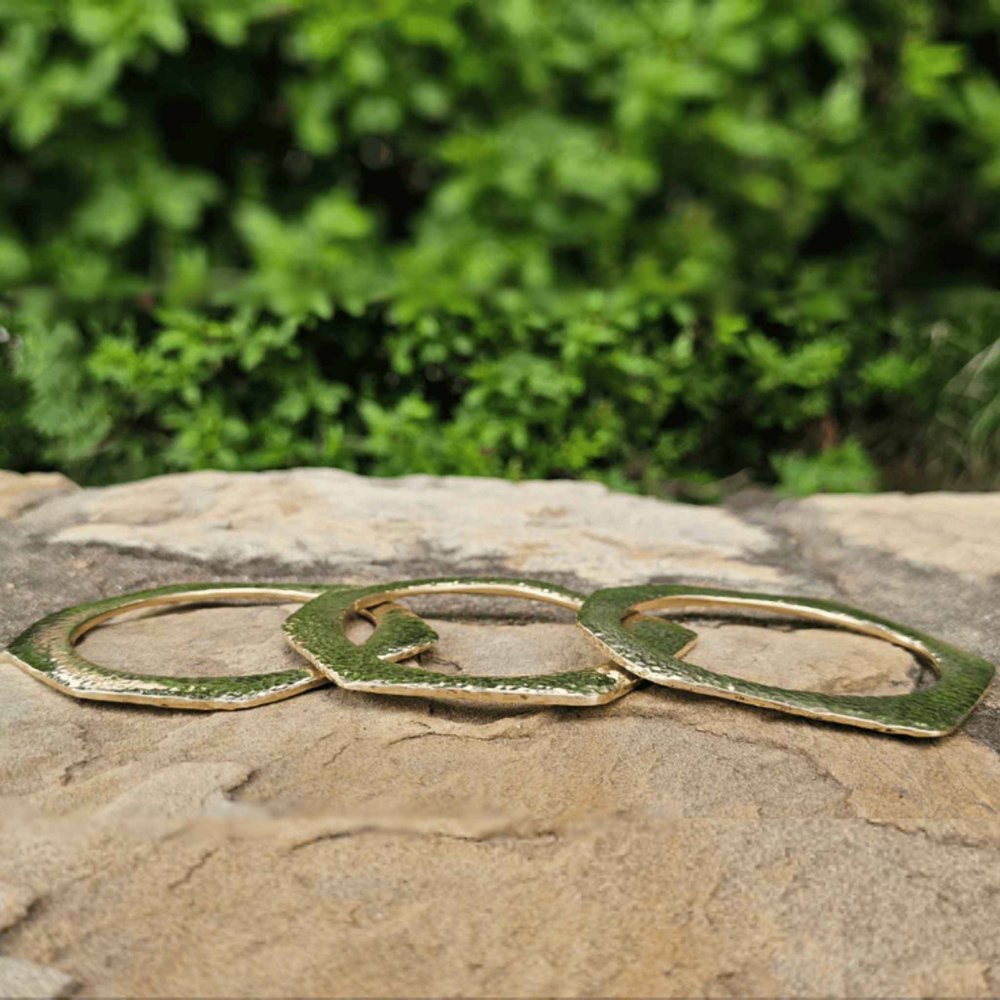 Three gold-toned metal wall hooks with a textured, hammered finish resting on a natural stone surface with a blurred green garden background. Shop Kamawe