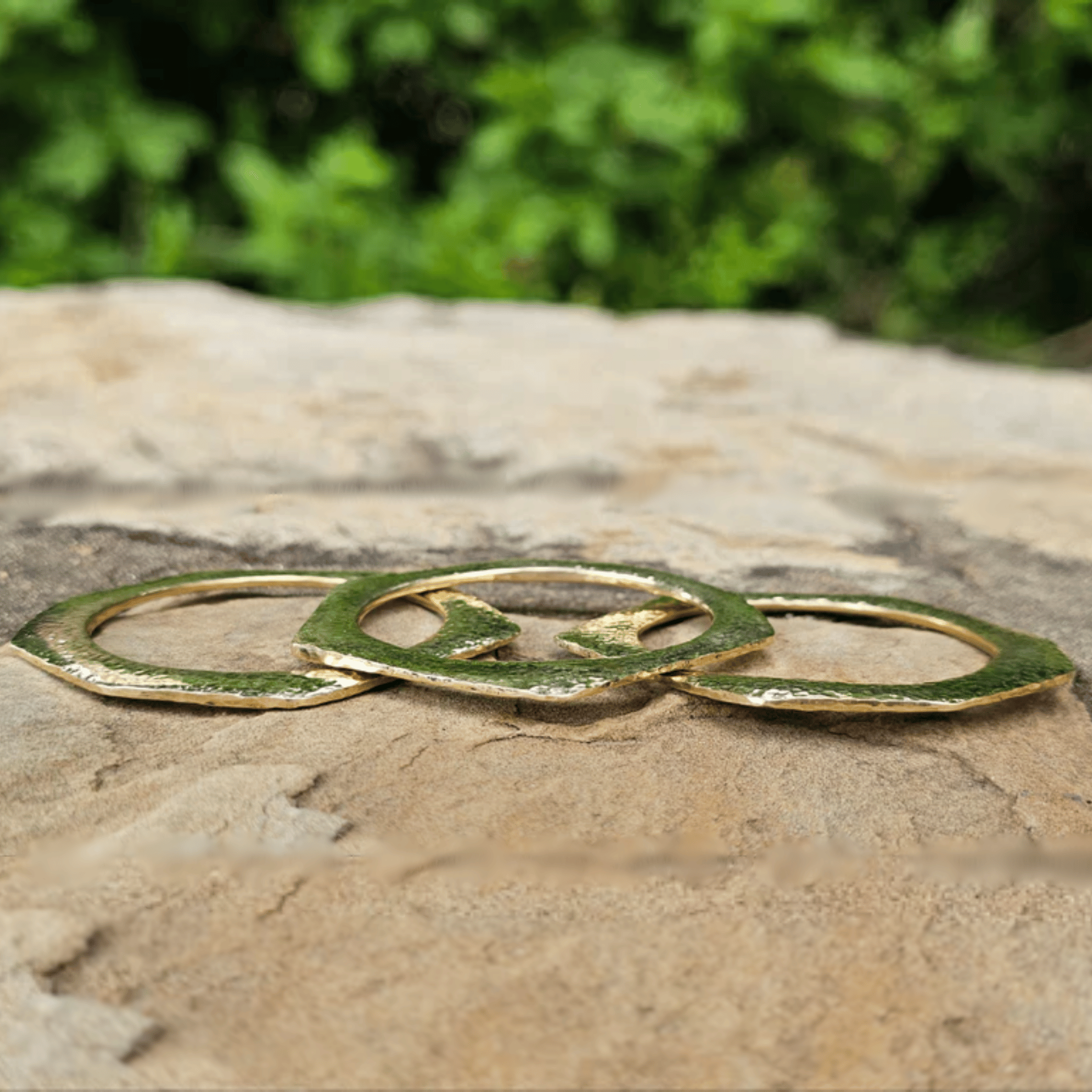 Three interlocking gold-toned metal rings with a textured green patina, resting on a rough stone surface with a blurred green foliage background. Shop Kamawe