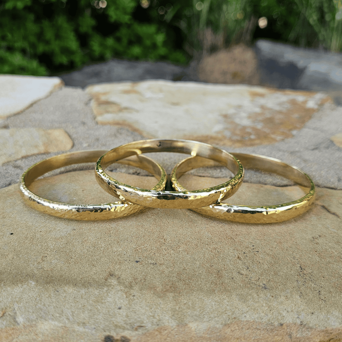 Three textured gold rings arranged in an overlapping circle on a natural stone surface with a blurred green garden background. Shop Kamawe