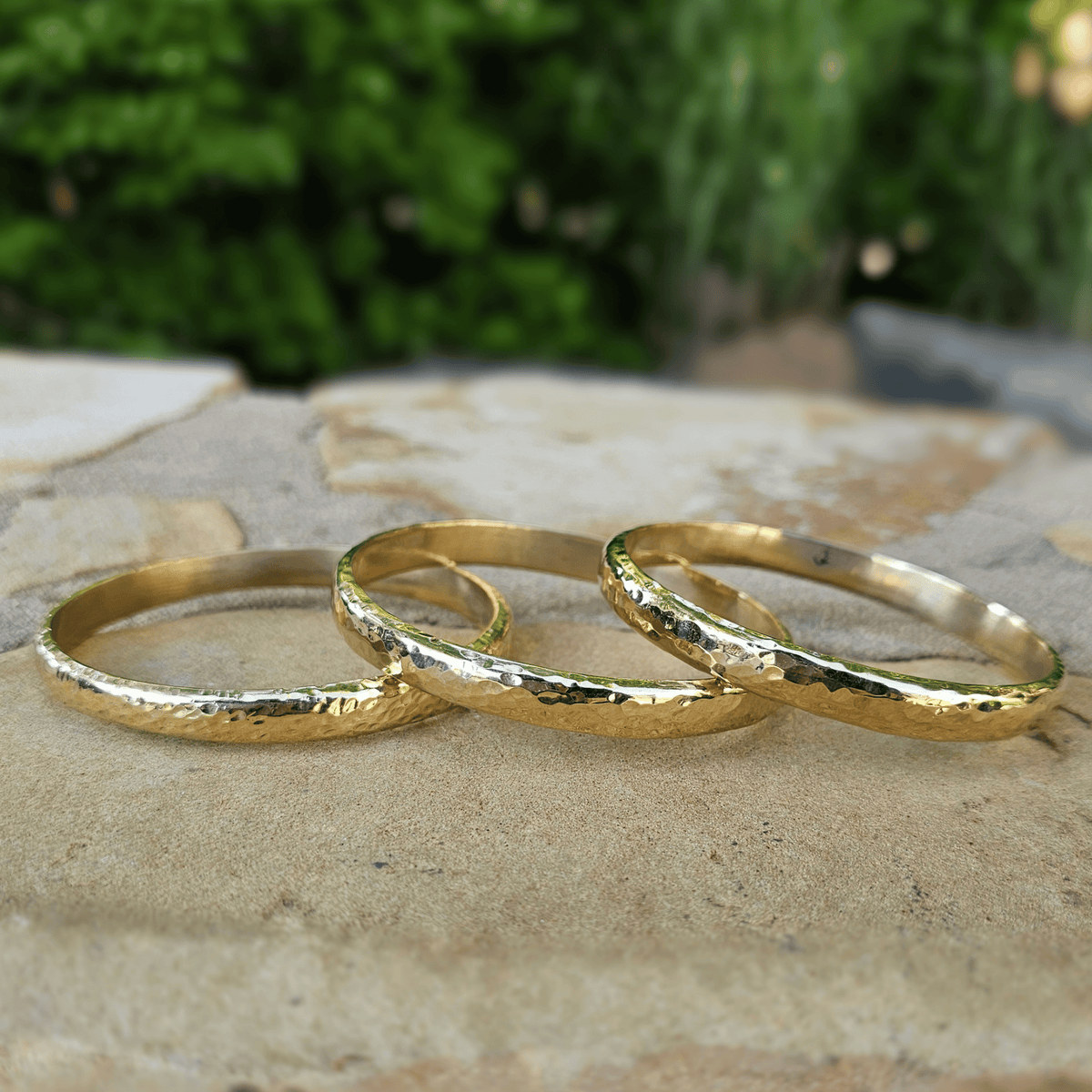 Three textured gold bands rings arranged on a stone surface with a blurred green outdoor background. Shop Kamawe