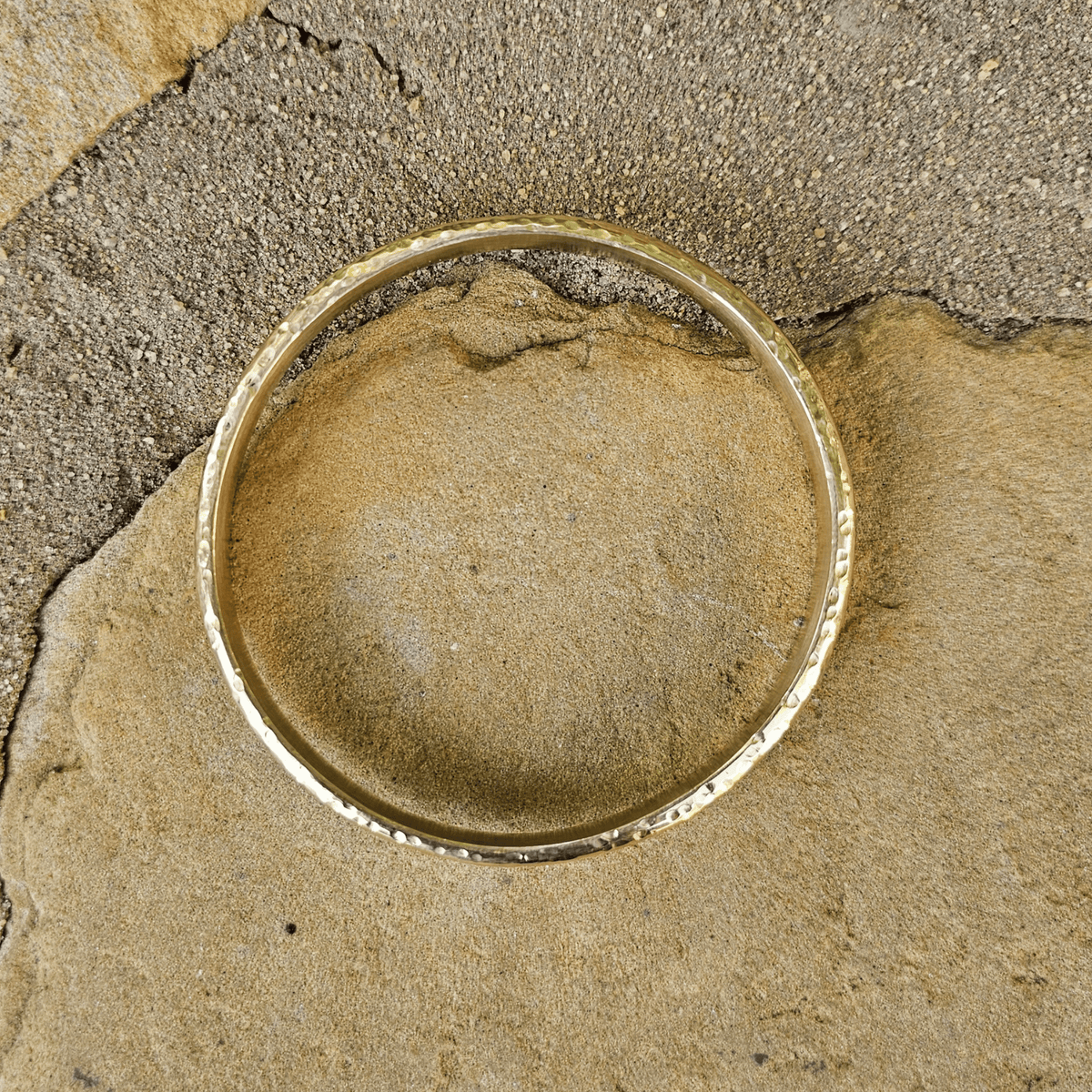 A hammered gold bangle bracelet resting on a textured stone surface. Shop Kamawe