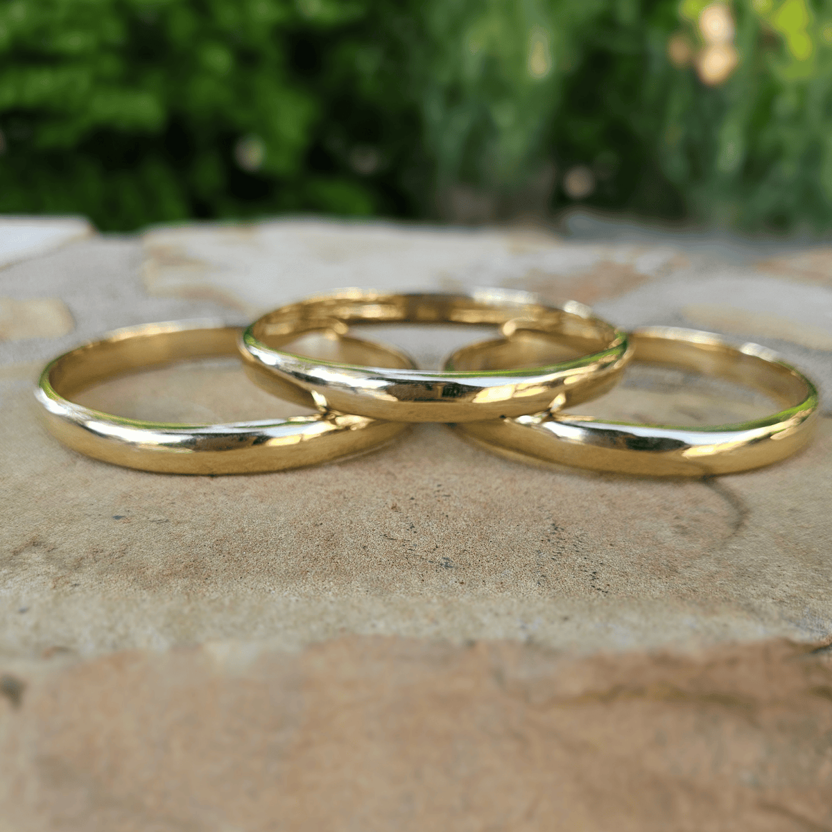 Three polished gold bracelets arranged in a circle on a textured stone surface with a blurred green garden background. Shop Kamawe