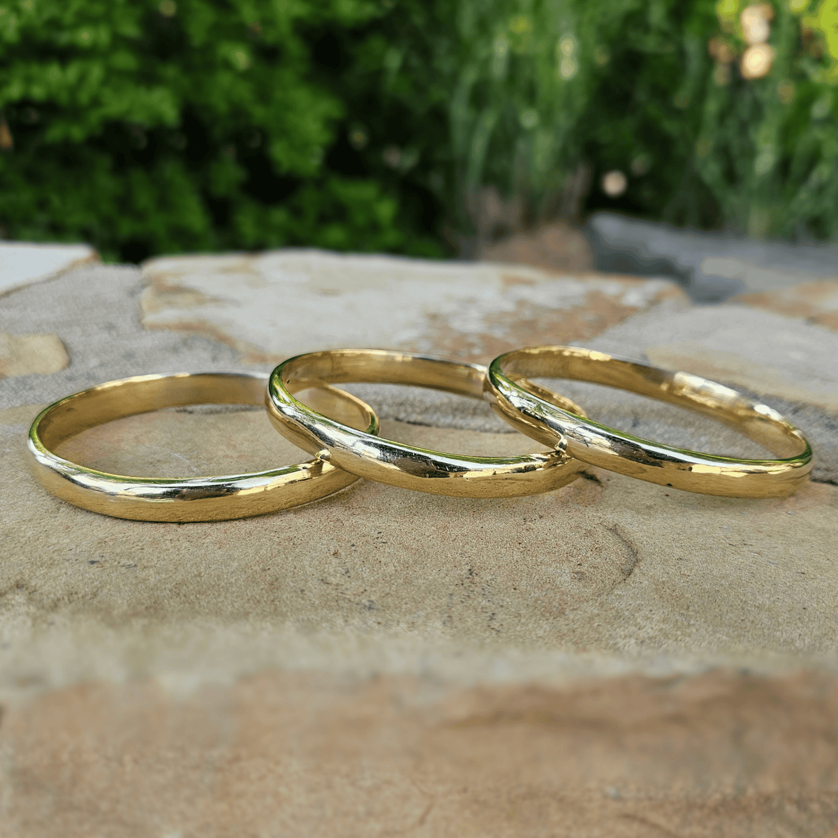 Three polished gold bracelets arranged in a circle on a textured stone surface with a blurred green garden background. Shop Kamawe