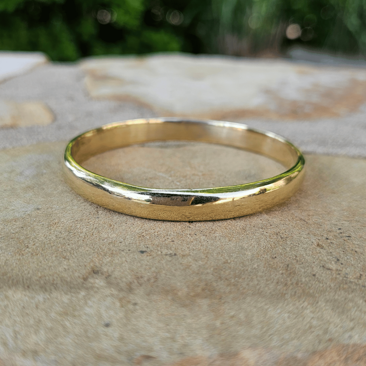 A simple polished gold band ring resting on a textured stone surface with a blurred green outdoor background. Shop Kamawe