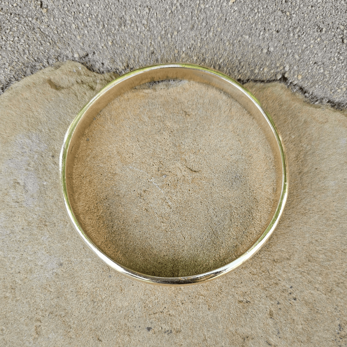 A polished gold bangle bracelet resting on a textured beige stone surface with a gray concrete background. Shop Kamawe