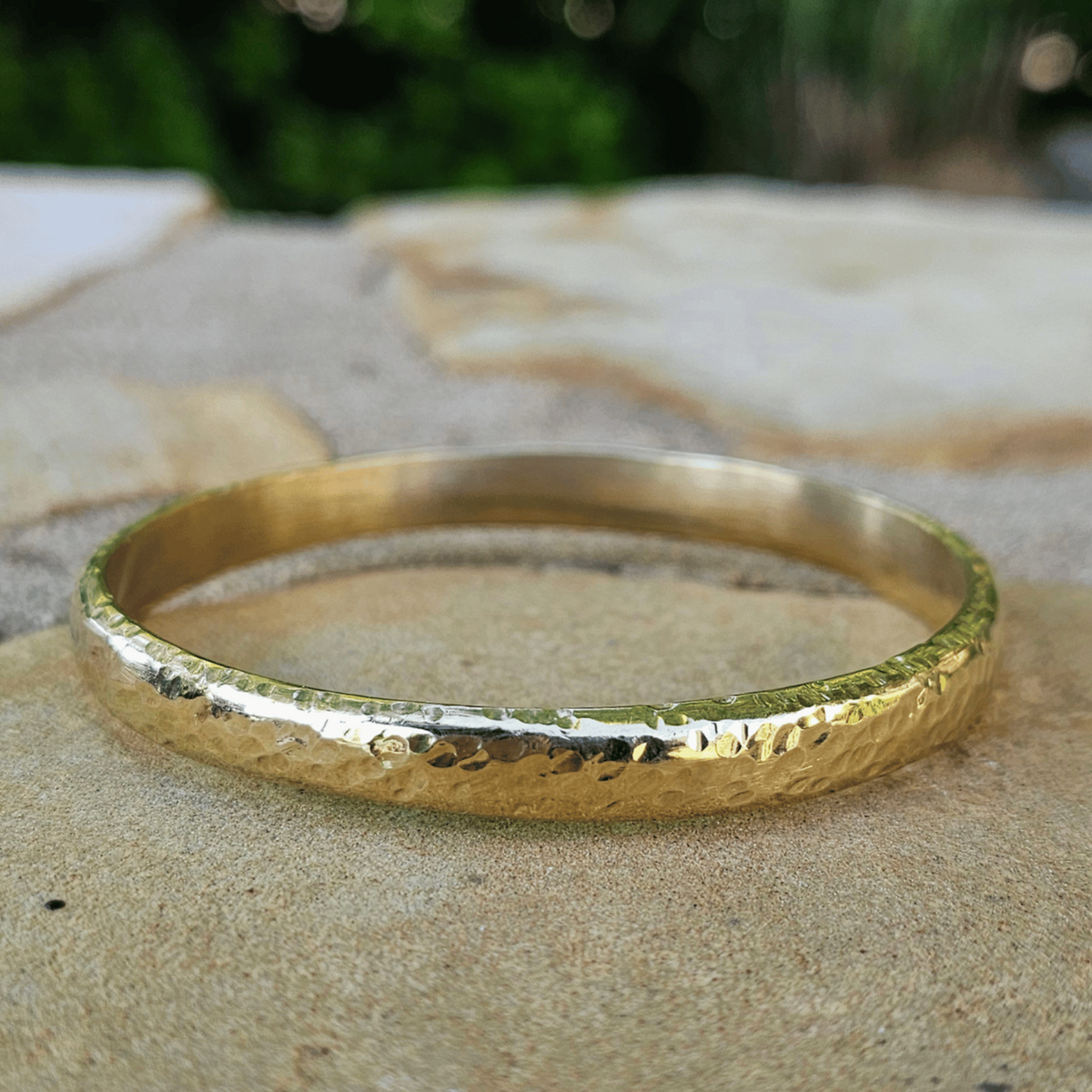 A hammered gold bangle bracelet resting on a textured stone surface with a blurred green outdoor background. Shop Kamawe