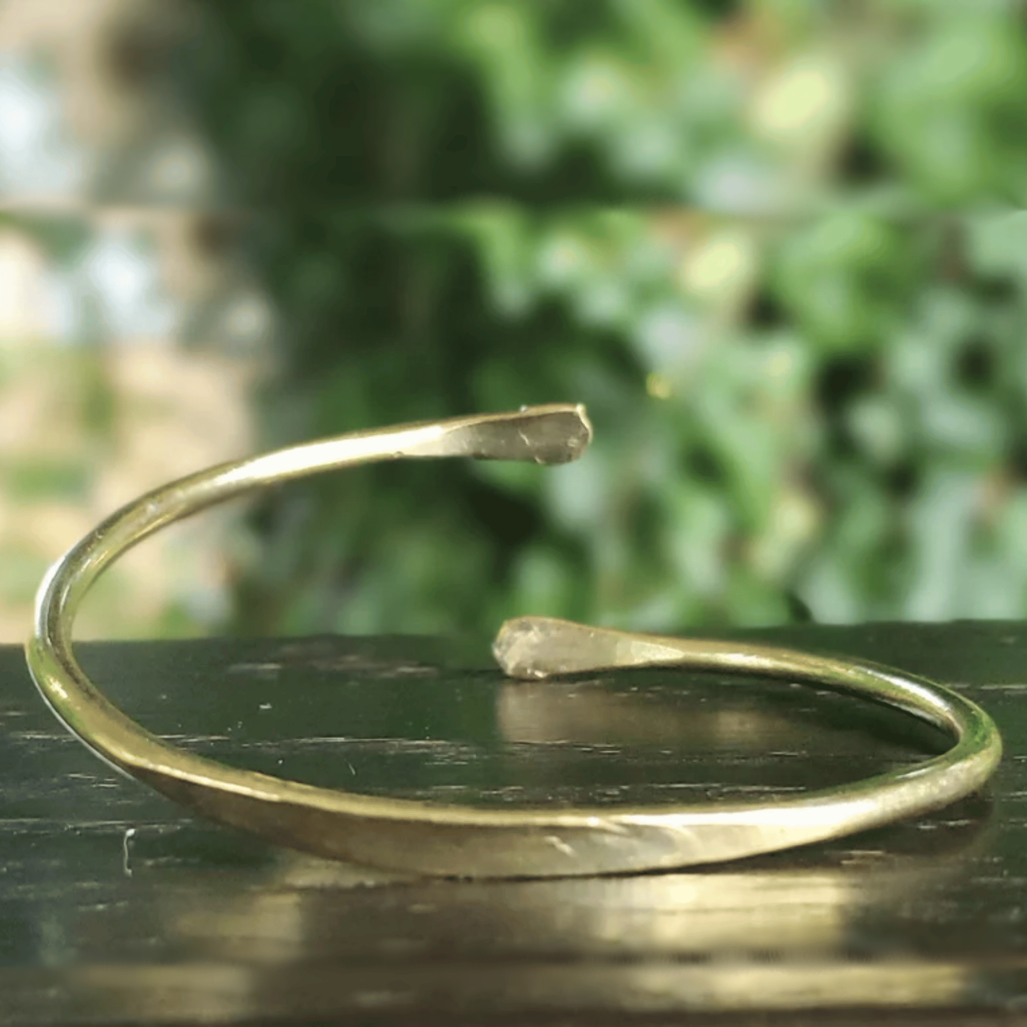 A hammered brass cuff bracelet with a rustic, open-ended design, resting on a dark wooden surface with a blurred green foliage background. Shop Kamawe