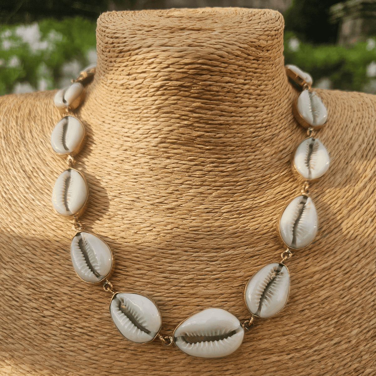 A cowrie shell necklace displayed on a woven beige mannequin bust. The necklace features multiple white cowrie shells with natural brown lines and small gold-toned metal links. Shop Kamawe