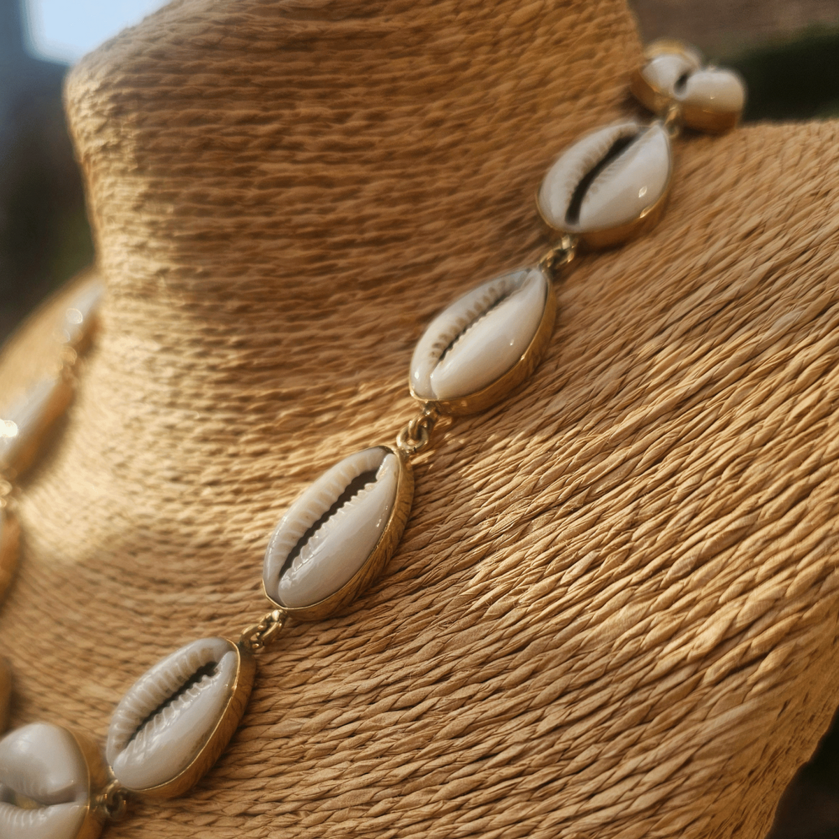 A cowrie shell necklace displayed on a woven beige mannequin bust. The necklace features multiple white cowrie shells with natural brown lines and small gold-toned metal links. Shop Kamawe