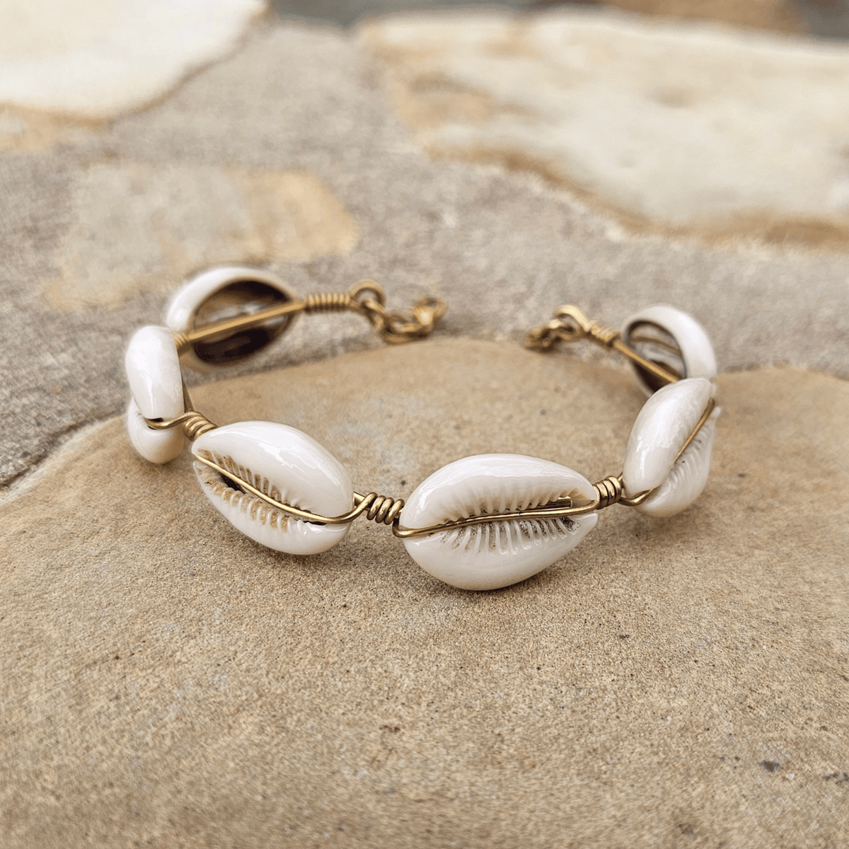 A handmade shell bracelet featuring five white cowrie shells wrapped in gold-toned wire, resting on a textured stone surface. Shop Kamawe