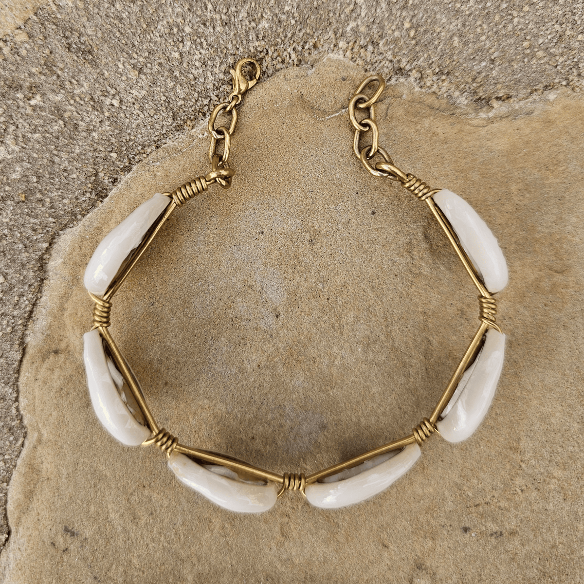 A handmade-style gold wire bracelet with white cowrie shells, displayed on a textured beige stone surface. Shop Kamawe