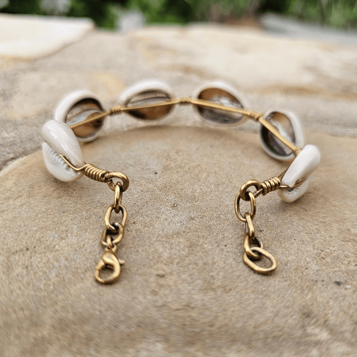 A handmade gold wire bracelet decorated with white cowrie shells and featuring adjustable lobster clasp chains, displayed on a textured stone surface. Shop Kamawe