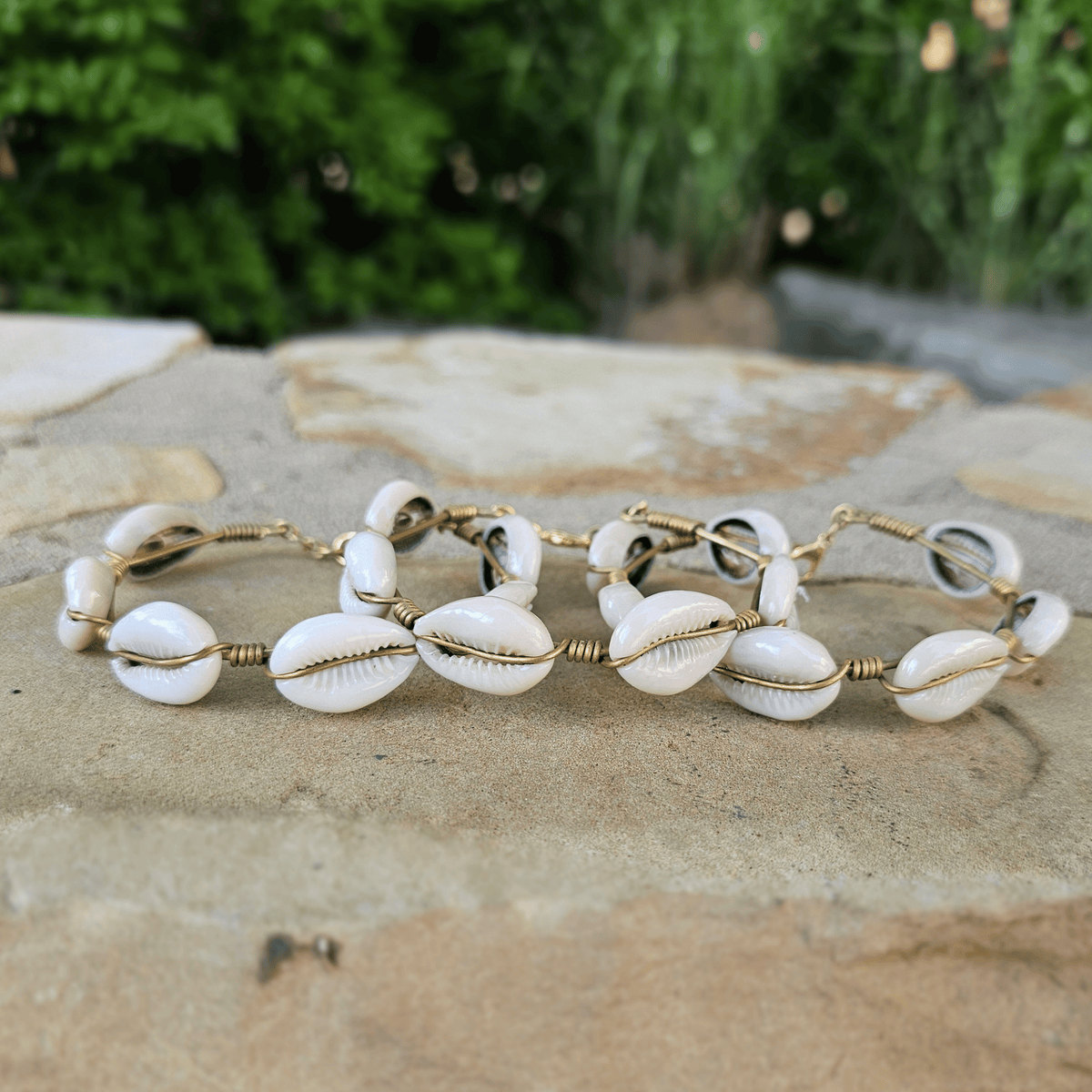 Three gold wire bracelets with white cowrie shells, arranged on a stone surface with a blurred green garden background. Shop Kamawe