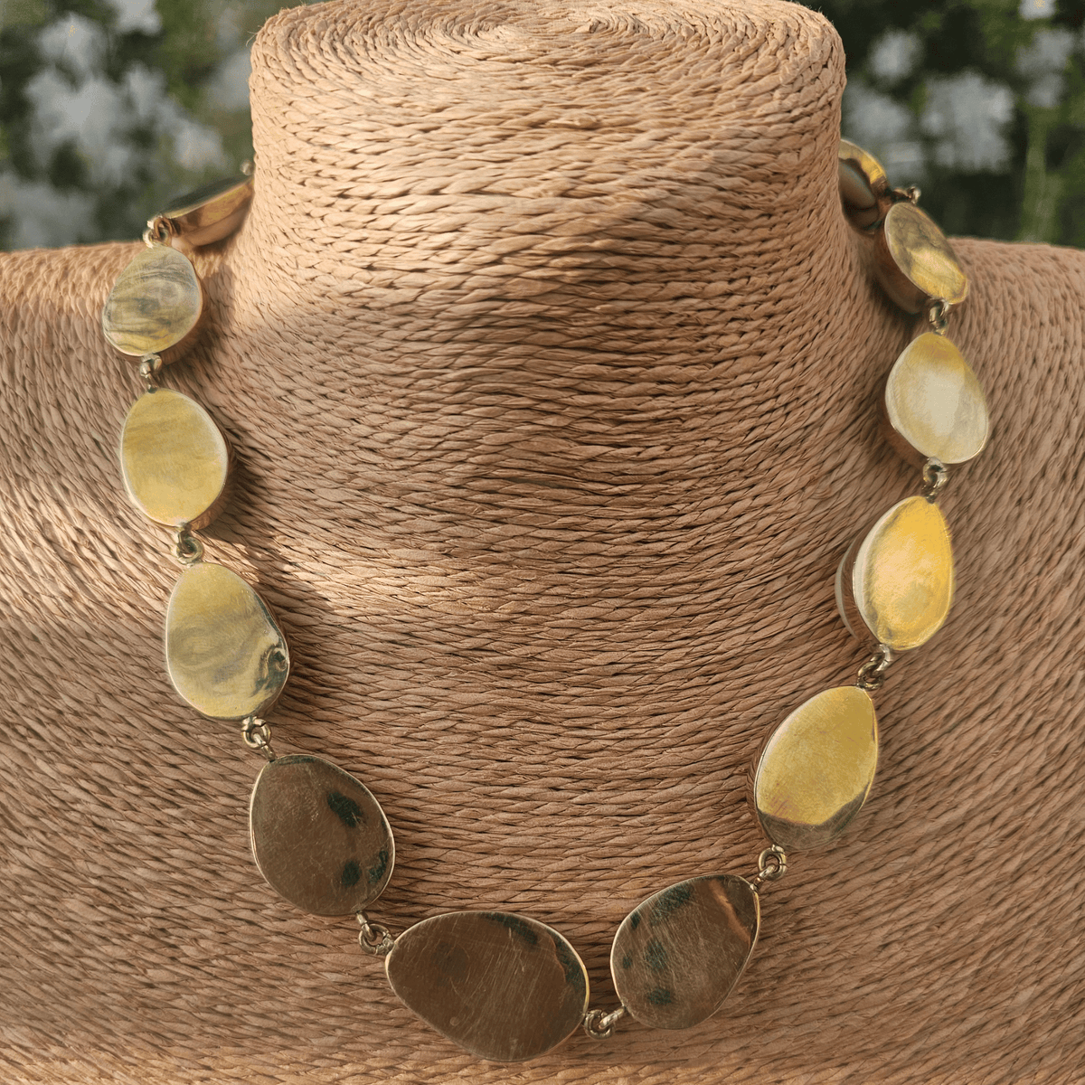 A cowrie shell necklace displayed on a woven beige mannequin bust. The necklace features multiple white cowrie shells with natural brown lines and small gold-toned metal links. Shop Kamawe