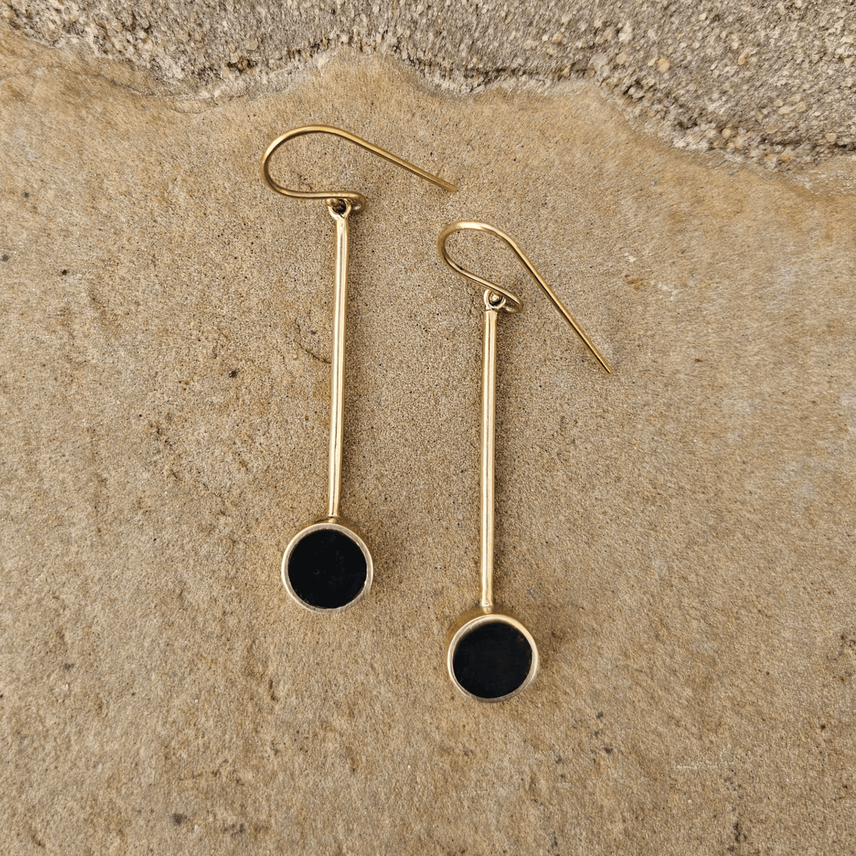 A pair of gold drop earrings with circular black inlay, displayed on a textured beige stone surface. Shop Kamawe
