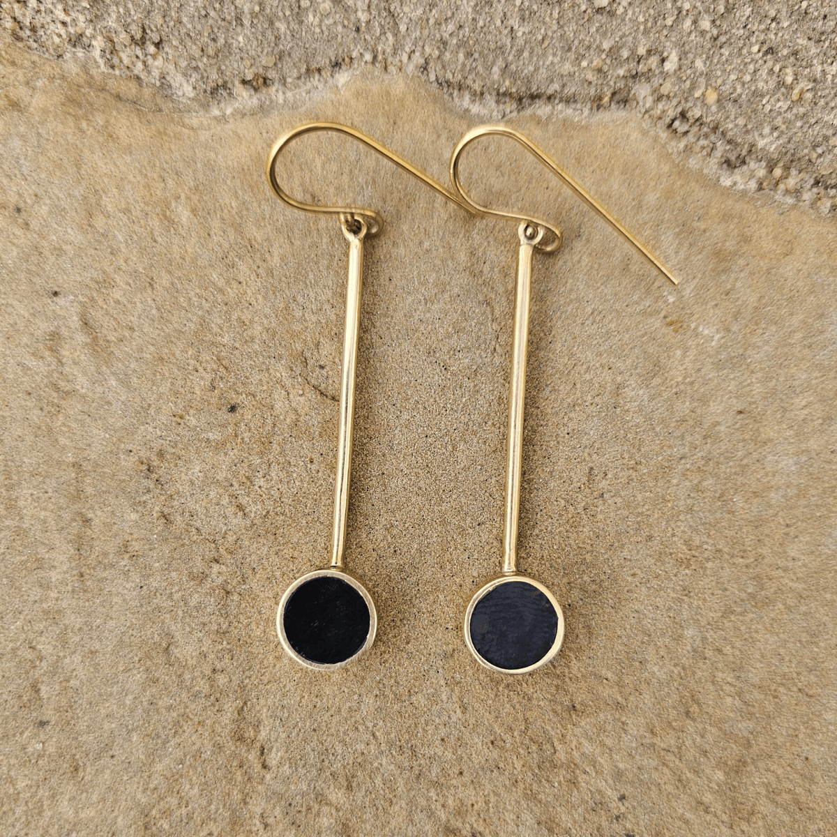 A pair of gold drop earrings with circular black inlay, displayed on a textured beige stone surface. Shop Kamawe