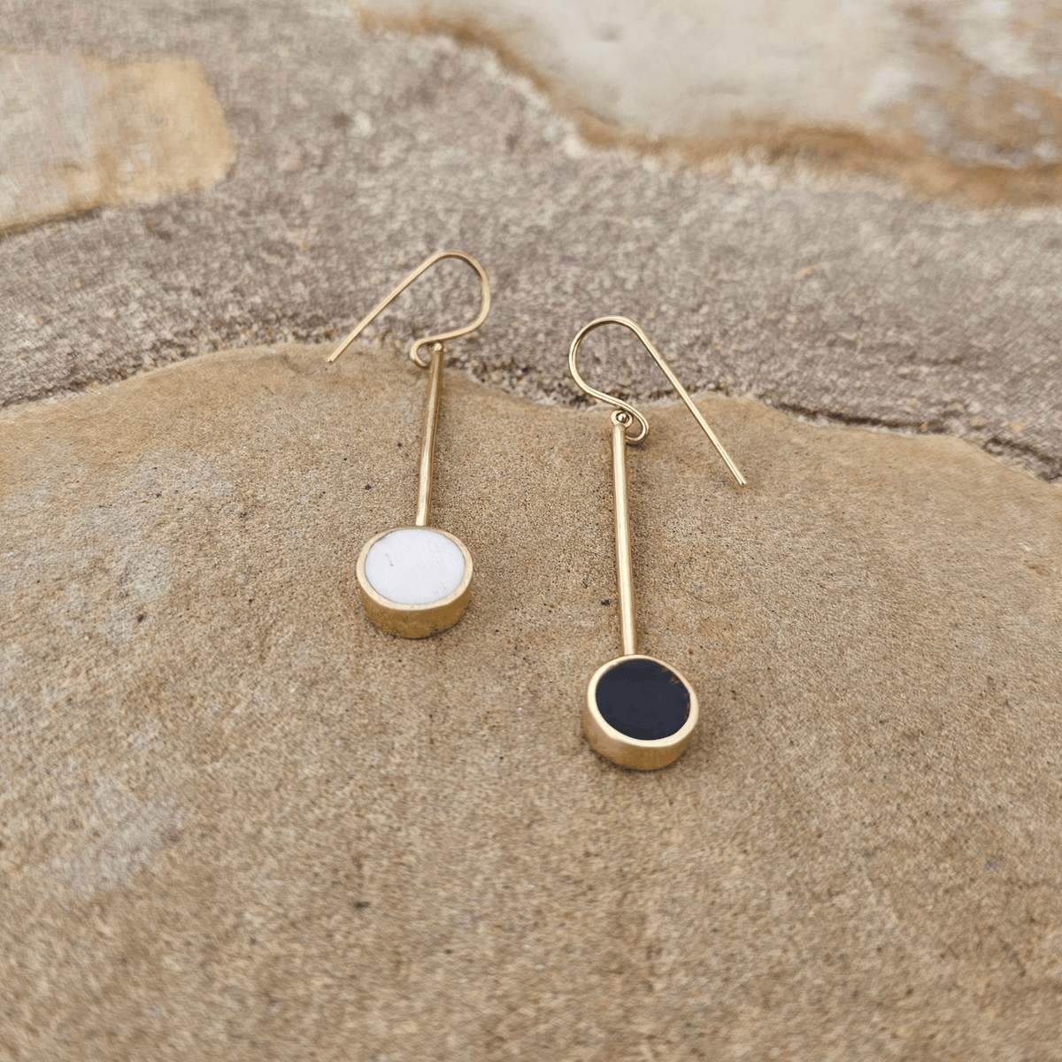 A pair of gold drop earrings with circular disc pendants, one white and one black, displayed on a textured beige stone surface. Shop Kamawe