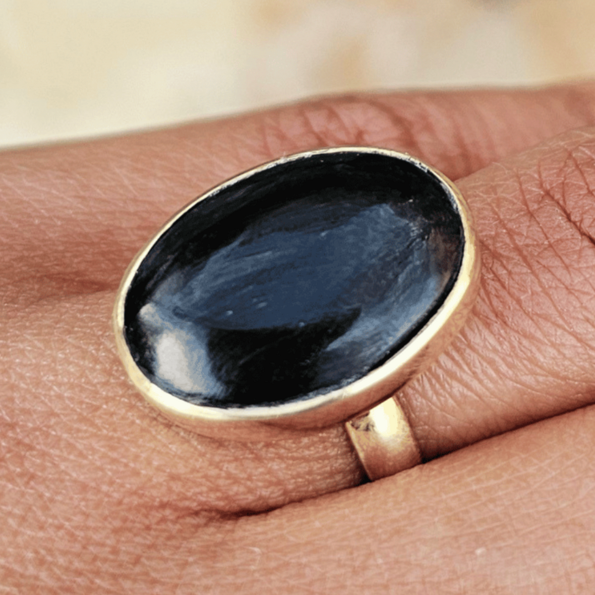 A gold ring with a large oval black cabochon gemstone worn on a person's finger, set against a blurred outdoor background. Shop Kamawe