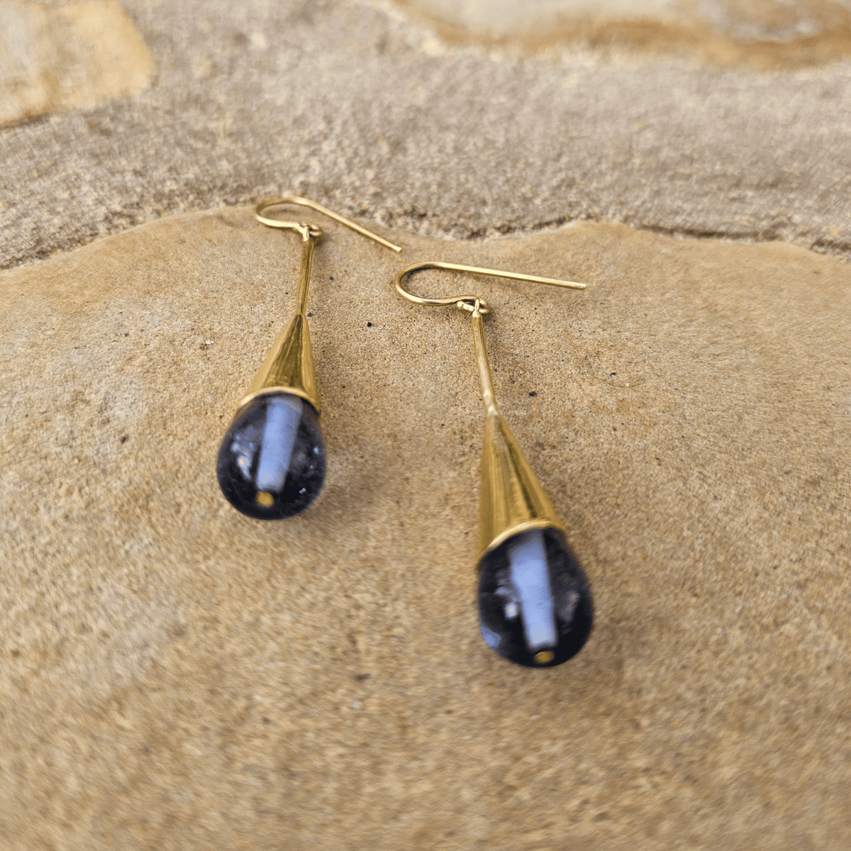 A pair of gold drop earrings with teardrop-shaped blue gemstone pendants, displayed on a textured tan stone surface. Shop Kamawe
