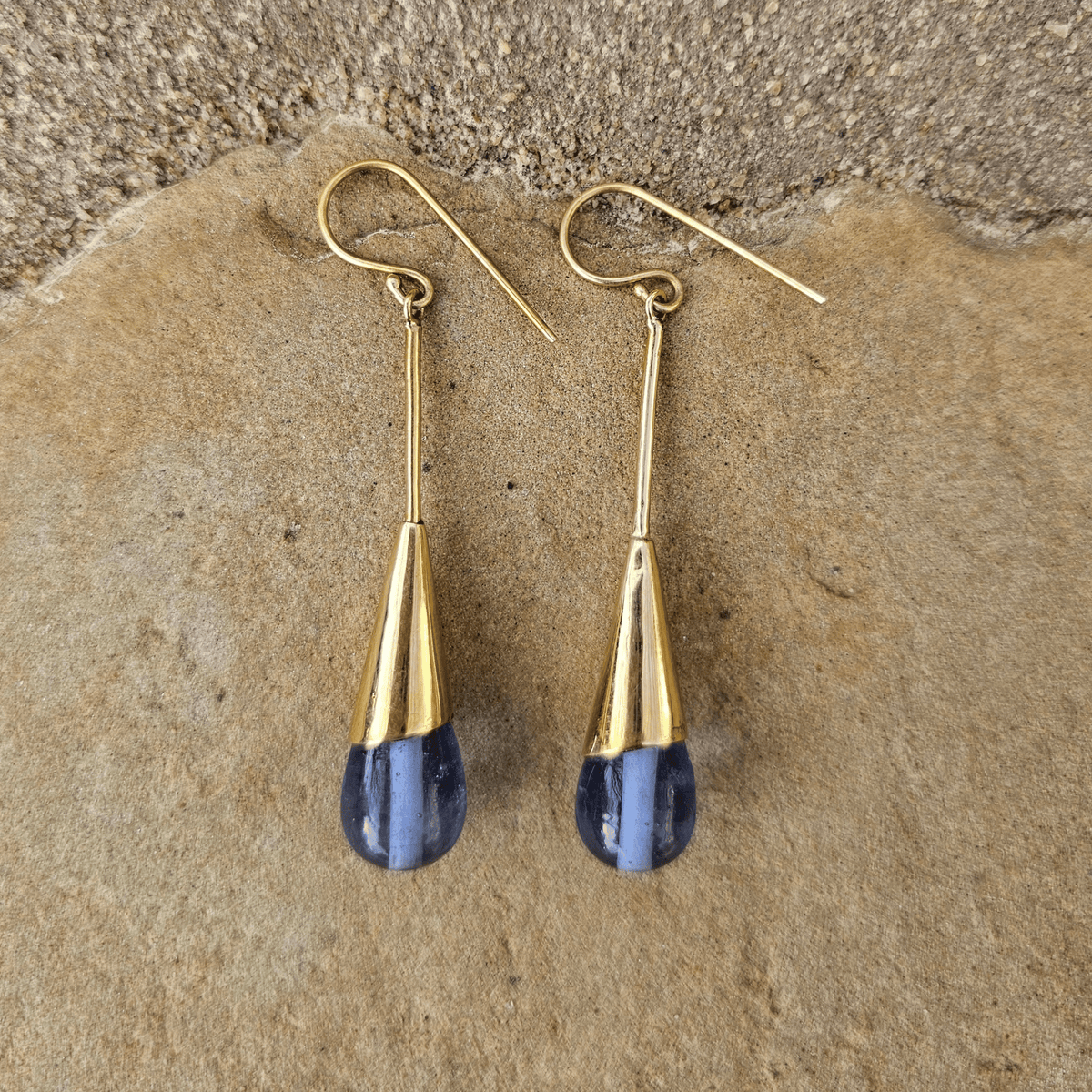 A pair of gold drop earrings with teardrop-shaped blue gemstone pendants, displayed on a textured tan stone surface. Shop Kamawe