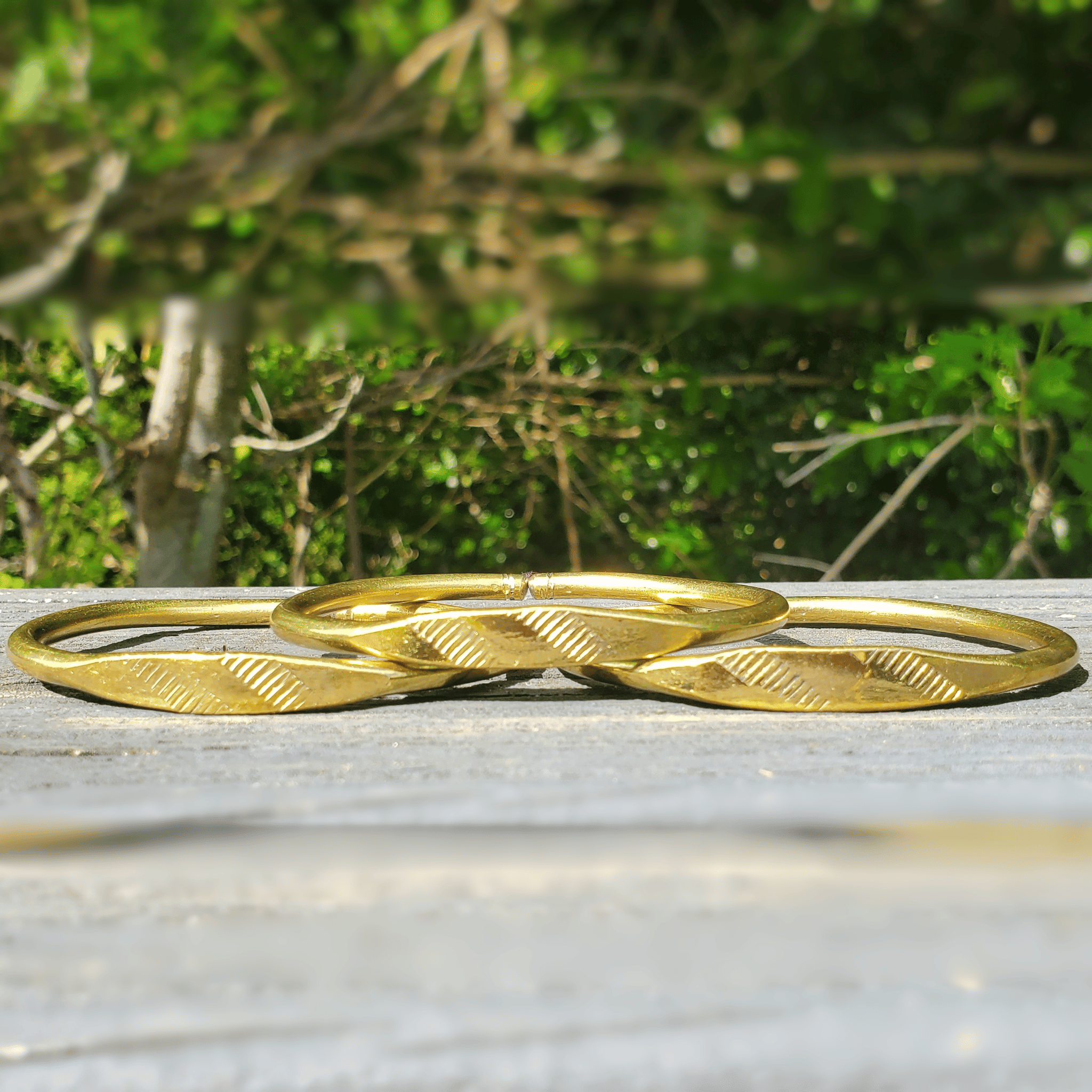 Three gold-toned bangles with a textured, ribbed leaf-like pattern resting on a weathered gray wooden surface with a blurred green outdoor background. Shop Kamawe