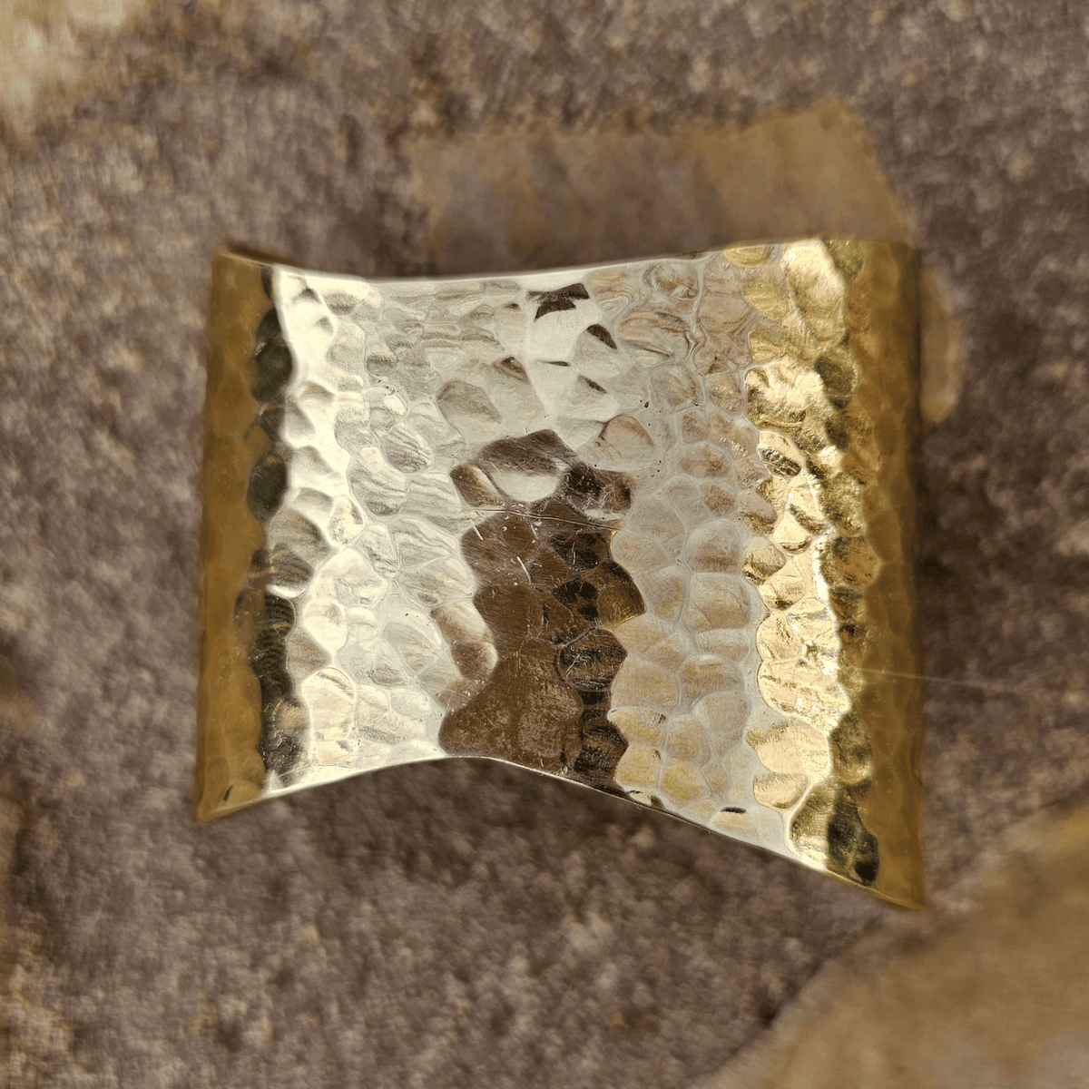 A hammered metal cuff bracelet with a textured, reflective surface, photographed on a natural stone background. Shop Kamawe