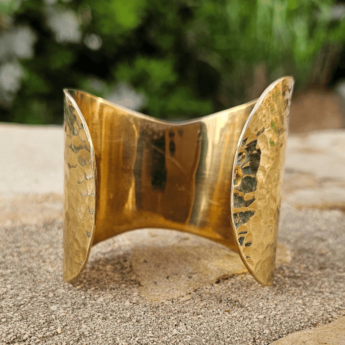 A hammered gold cuff bracelet standing upright on a sandy surface with a blurred green garden background. Shop Kamawe
