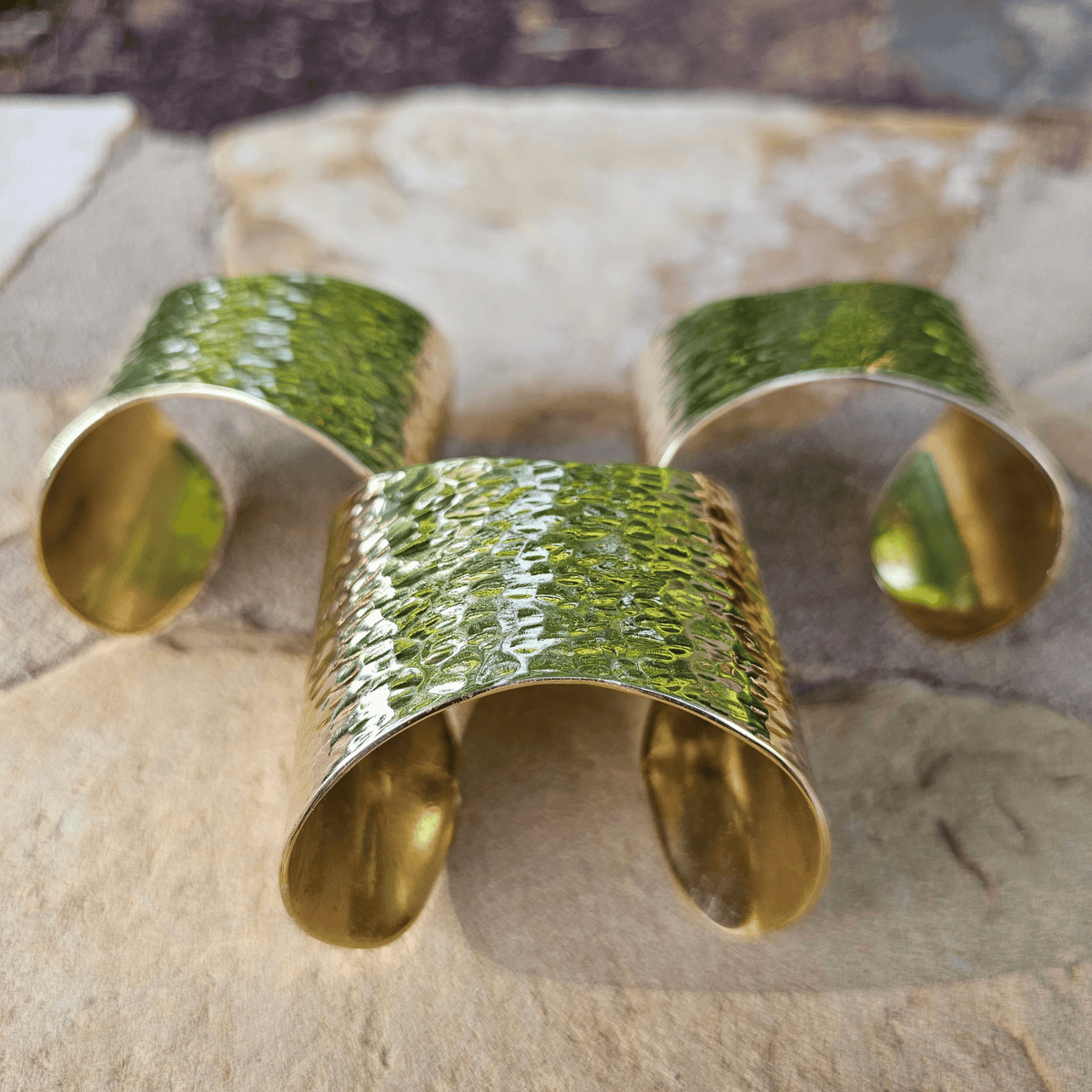 Three hammered gold cuff bracelets arranged on a light stone surface. Shop Kamawe