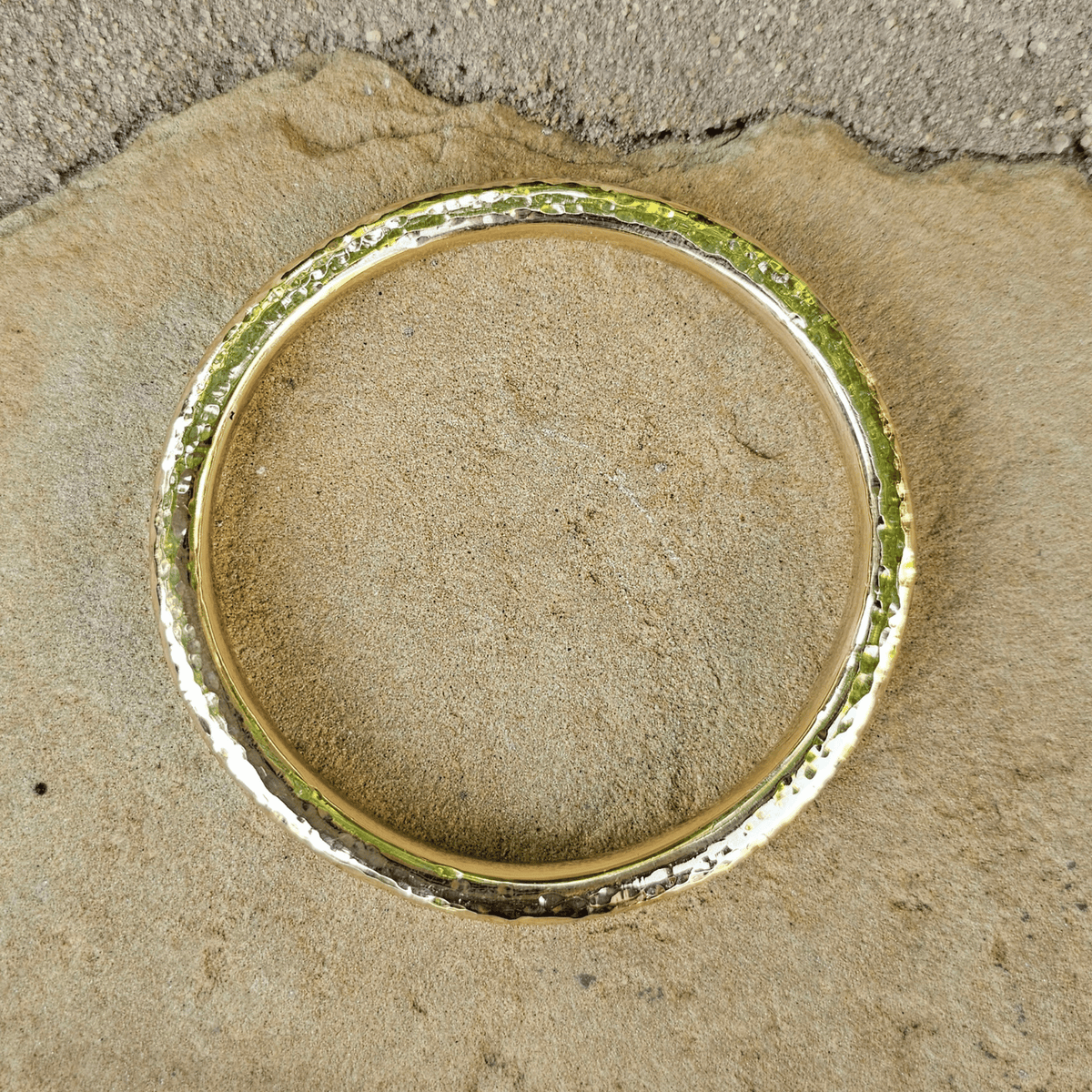 A hammered gold bangle bracelet resting on a textured beige stone surface. Shop Kamawe