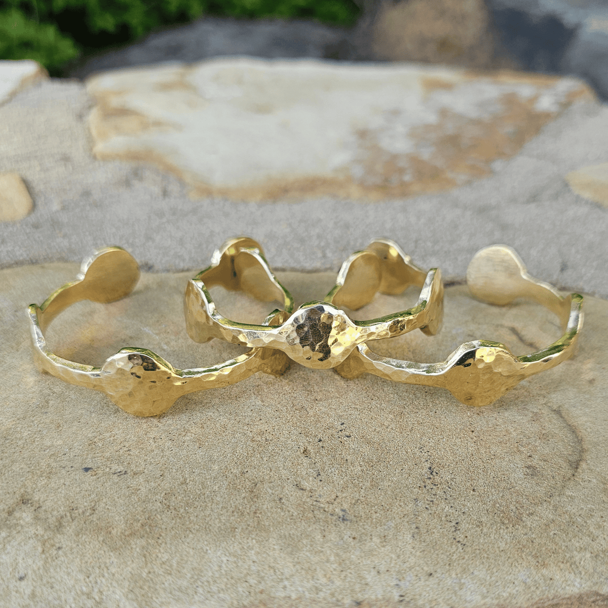 Three gold-toned textured open cuff bracelets arranged on a natural stone surface outdoors. Shop Kamawe