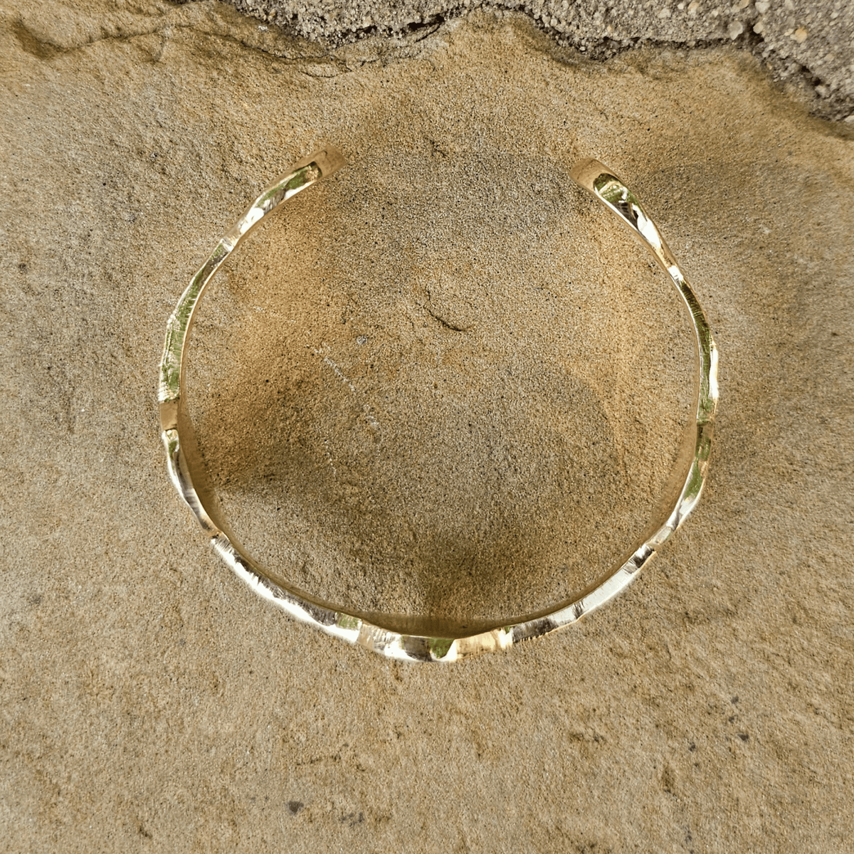 A hammered gold bangle bracelet displayed on a textured stone surface. Shop Kamawe