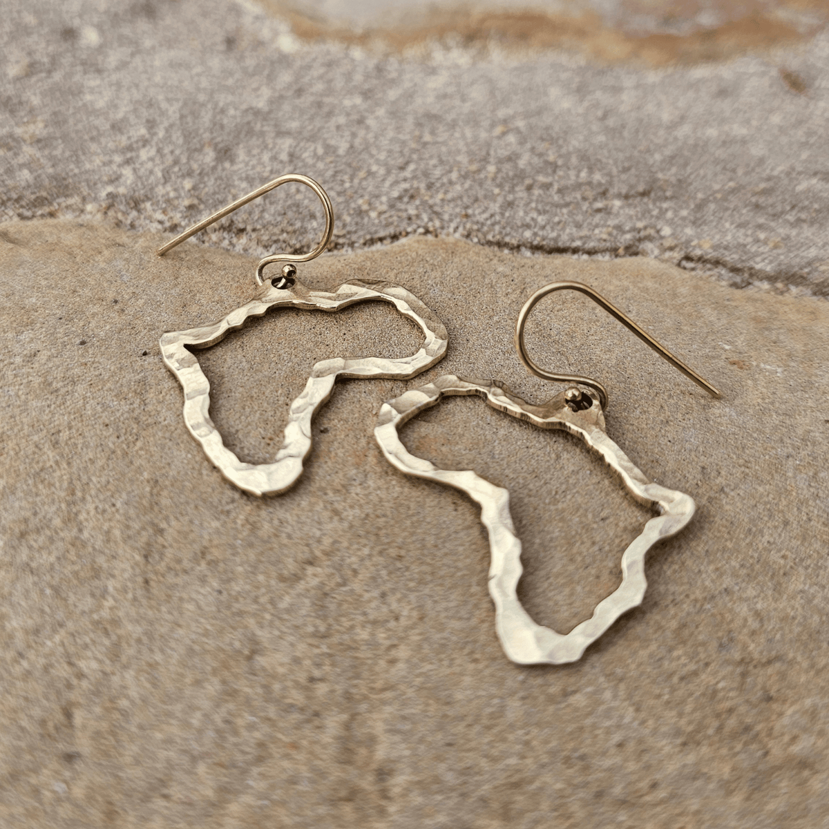 A pair of hammered metal earrings shaped like the outline of Africa, resting on a textured stone surface. Shop Kamawe