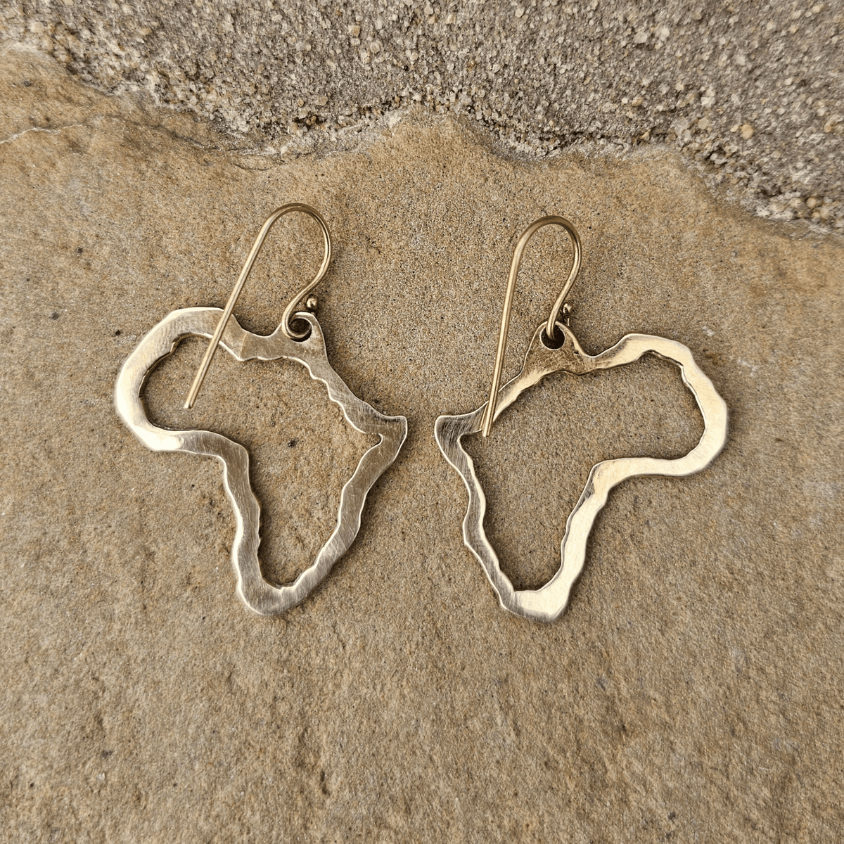A pair of dangling earrings shaped like the outline of Africa, made from textured silver-toned metal with fishhook wires, displayed on a natural stone surface. Shop Kamawe