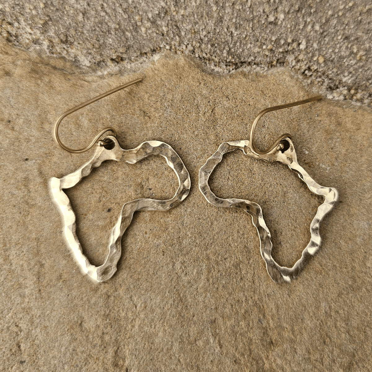 A pair of hammered metal earrings shaped like the outline of Africa, resting on a textured stone surface. Shop Kamawe