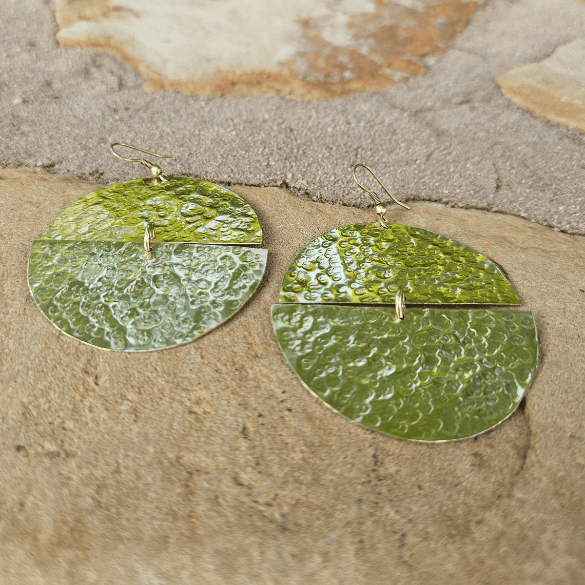 A pair of hammered green metal dangle earrings with a split circular design and small gold hooks, displayed on a textured beige stone surface. Shop Kamawe