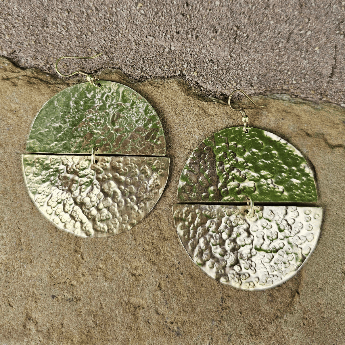 A pair of hammered green metal dangle earrings with a split circular design and small gold hooks, displayed on a textured beige stone surface. Shop Kamawe