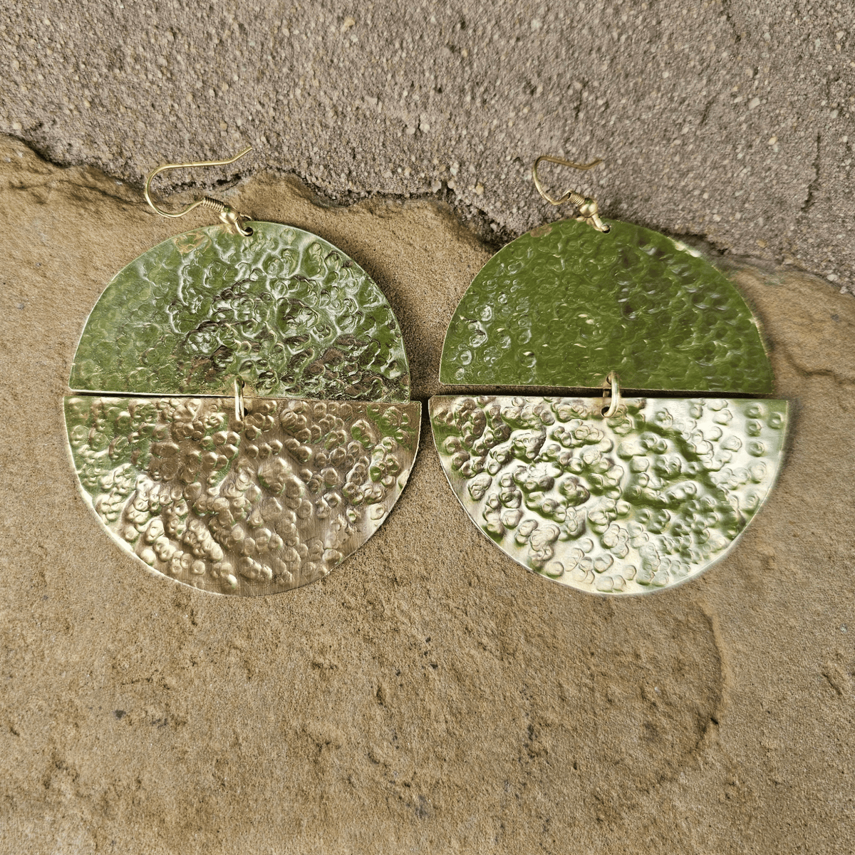 A pair of hammered green metal dangle earrings with a split circular design and small gold hooks, displayed on a textured beige stone surface. Shop Kamawe