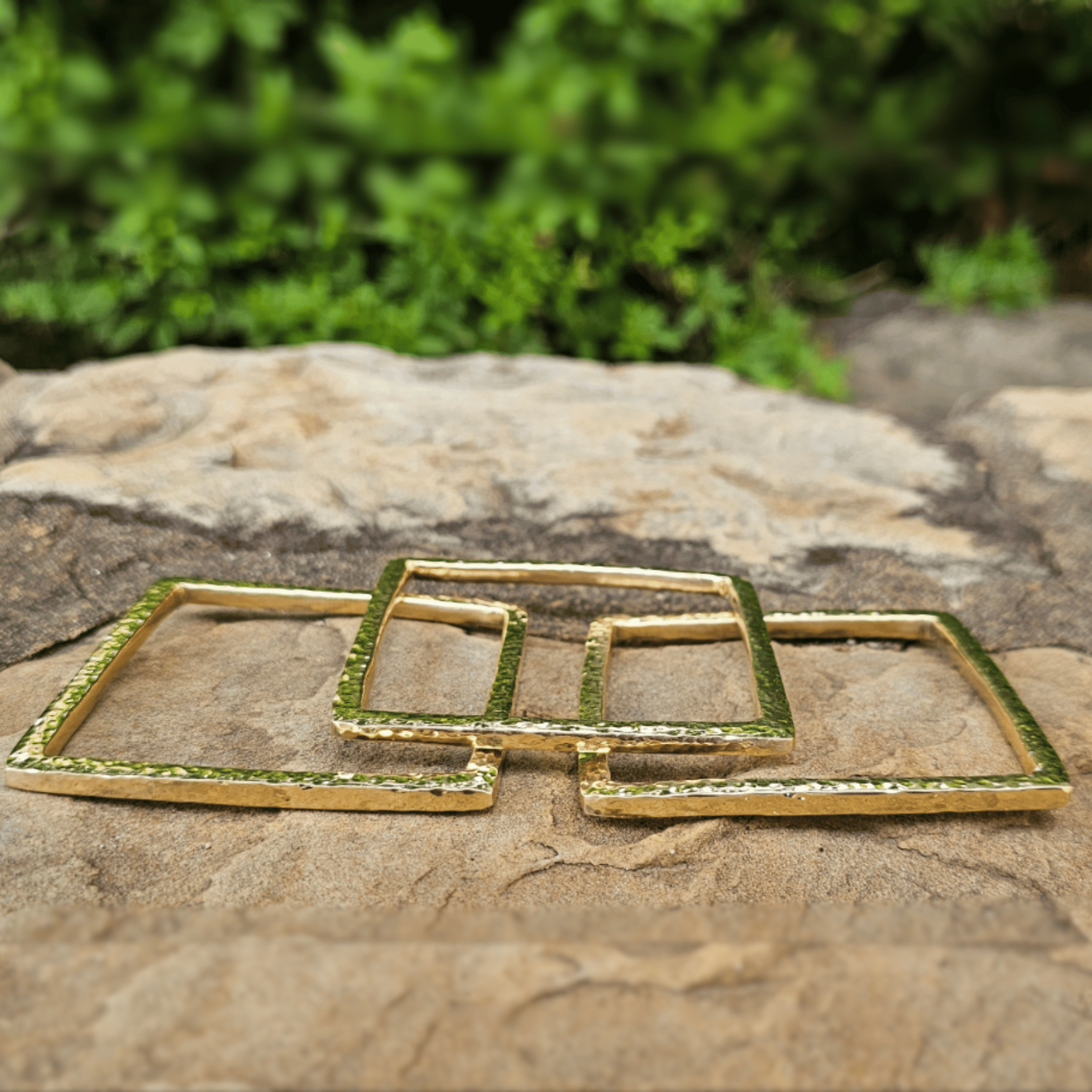 A gold-toned geometric square link chain earring resting on a textured stone surface with a blurred green garden background. Shop Kamawe