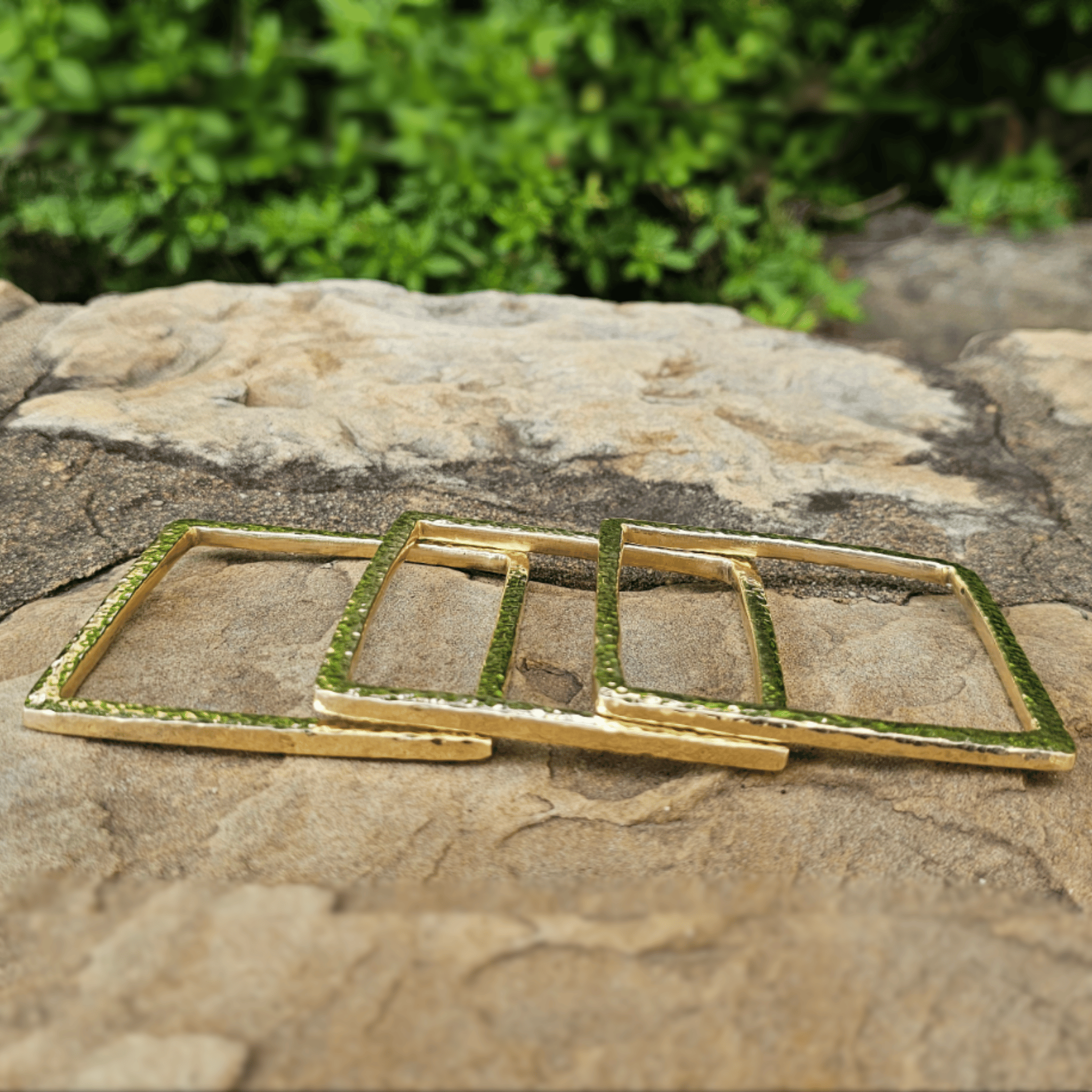 Three gold-toned square metal frames with a textured, hammered finish, arranged overlapping on a natural stone surface with green foliage in the background. Shop Kamawe