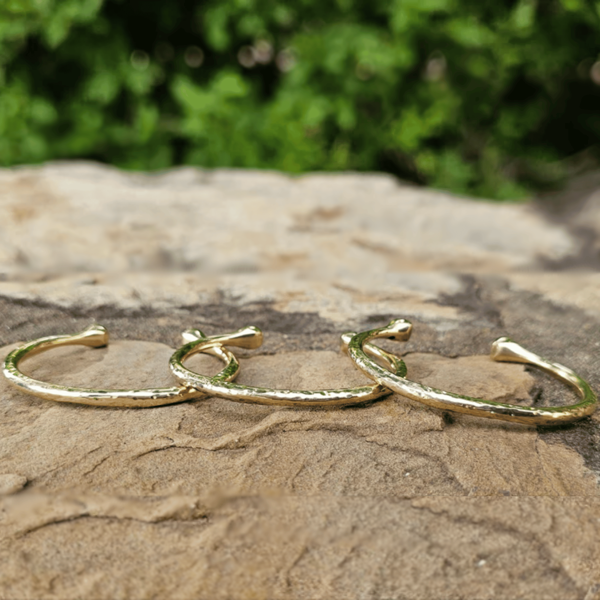 Three gold-toned textured open cuff earrings displayed on a natural stone surface with a blurred green outdoor background. Shop kamawe