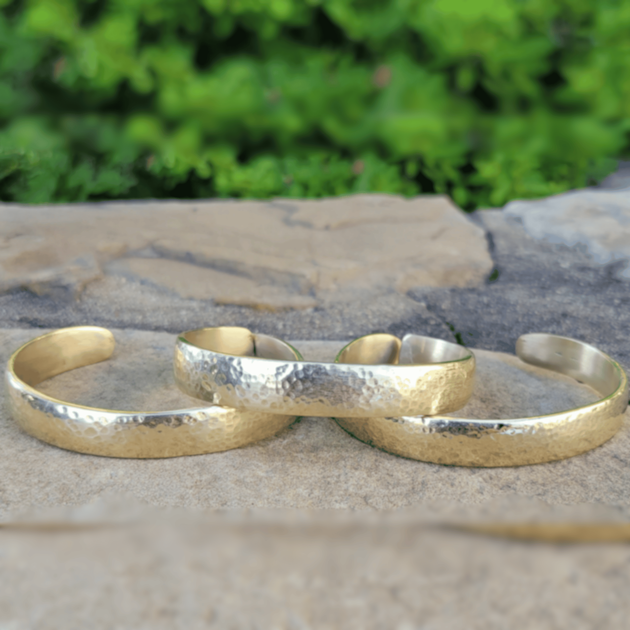Three hammered gold-tone cuff bracelets arranged on a stone surface with a blurred green outdoor background. Shop Kamawe