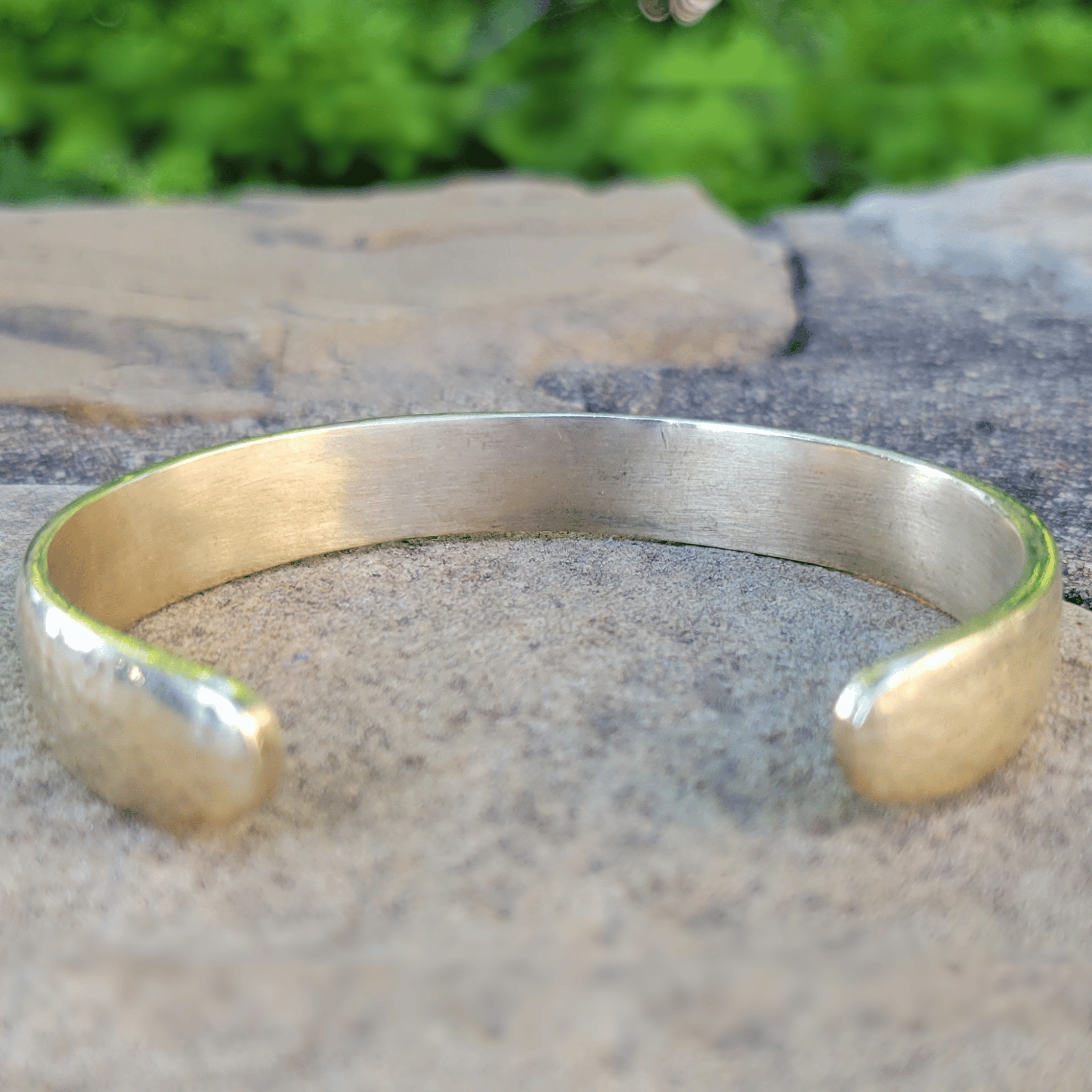 A hammered open cuff bracelet in a warm yellow-gold metal, resting on a textured stone surface with a blurred green background. Shop Kamawe