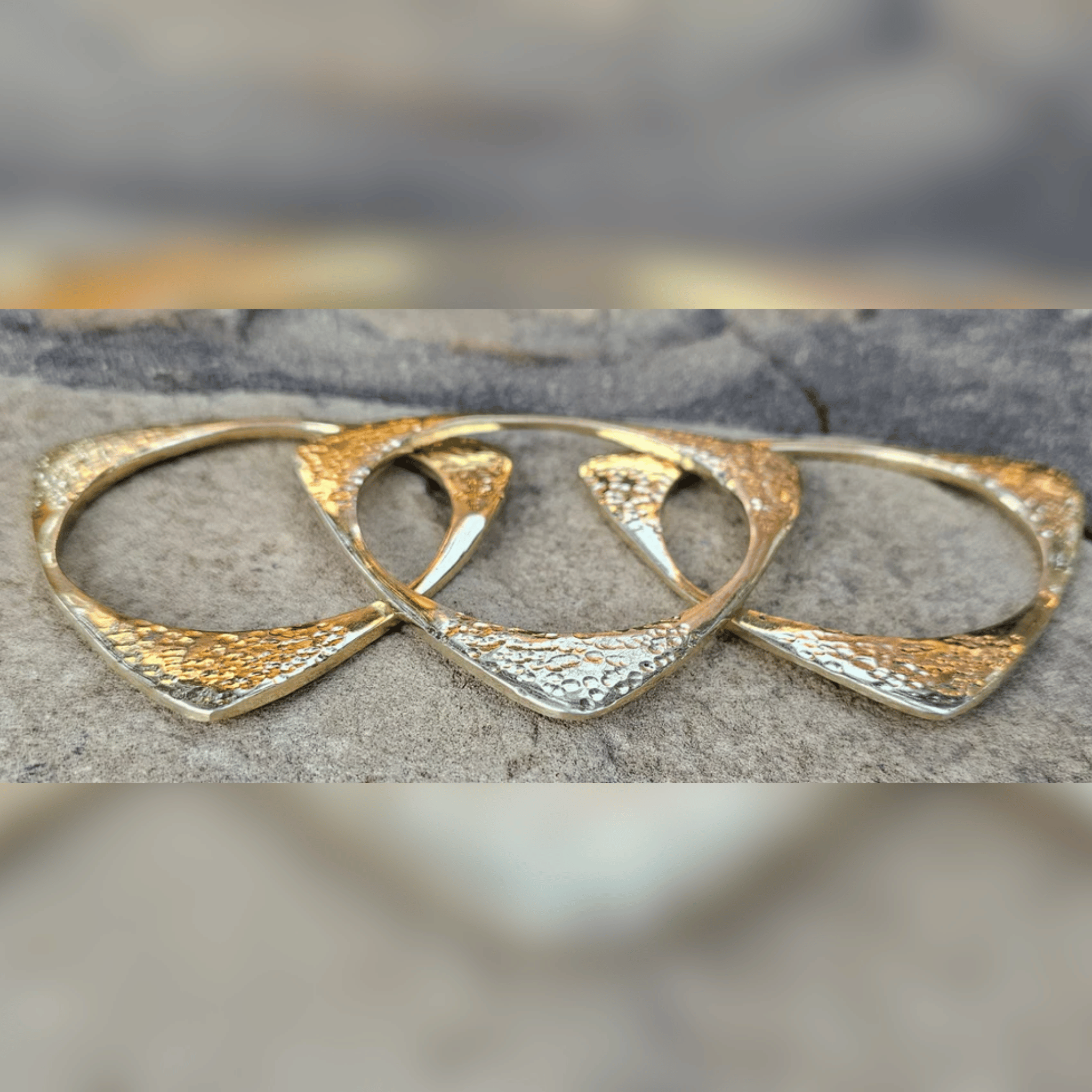 A pair of textured gold and silver geometric hoop earrings resting on a gray stone surface. Shop Kamawe