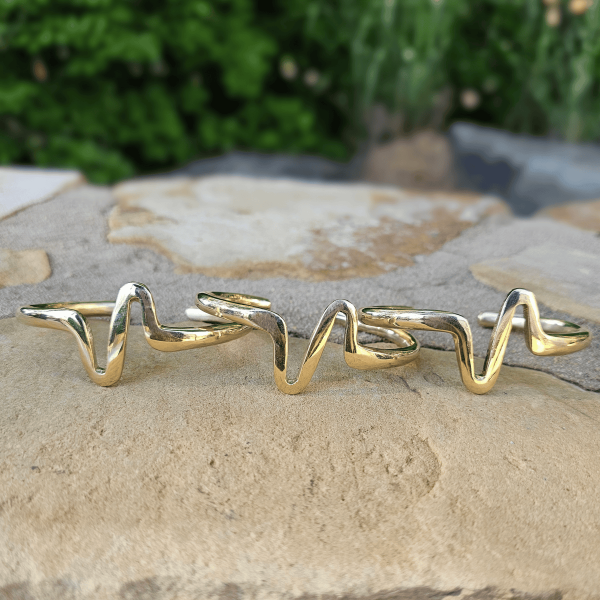 A gold-toned wavy ring with a unique zigzag design, displayed on a light beige stone surface with a blurred green garden background. Shop Kamawe