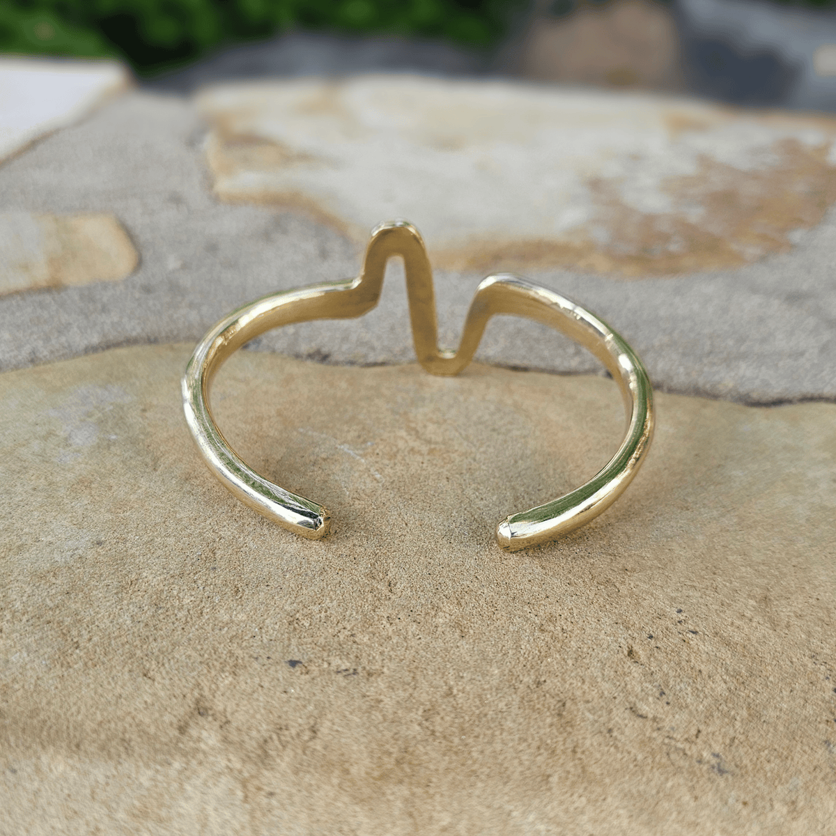A gold-toned open cuff ring with a small central peak, photographed resting on a textured beige stone surface outdoors. Shop Kamawe