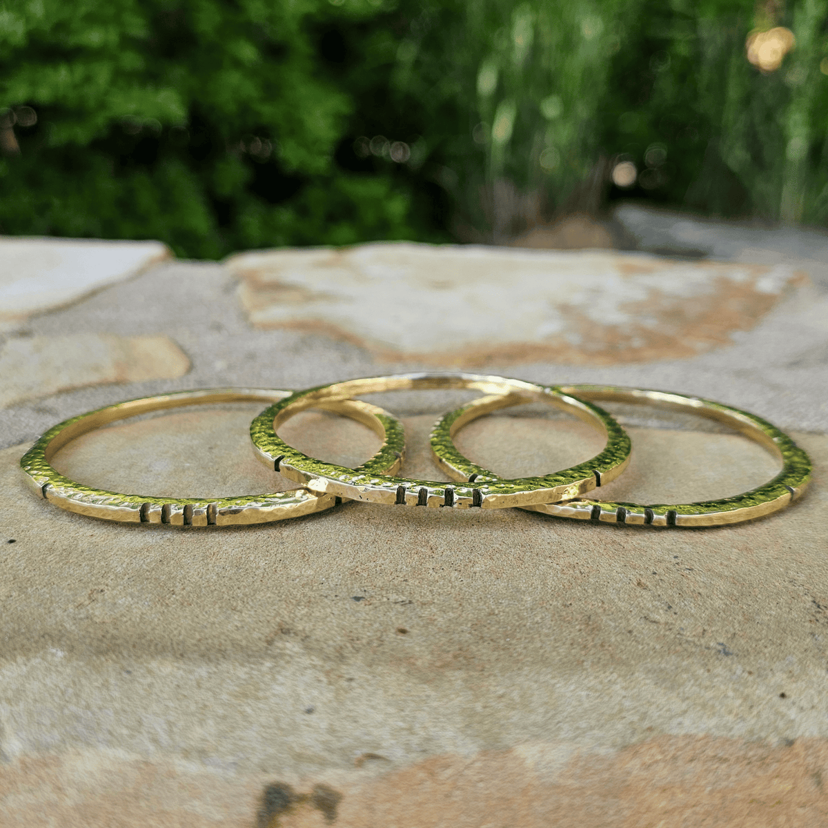 Three gold-toned textured bangles with small black stone accents, arranged on a stone surface with a blurred green outdoor background. Shop Kamawe