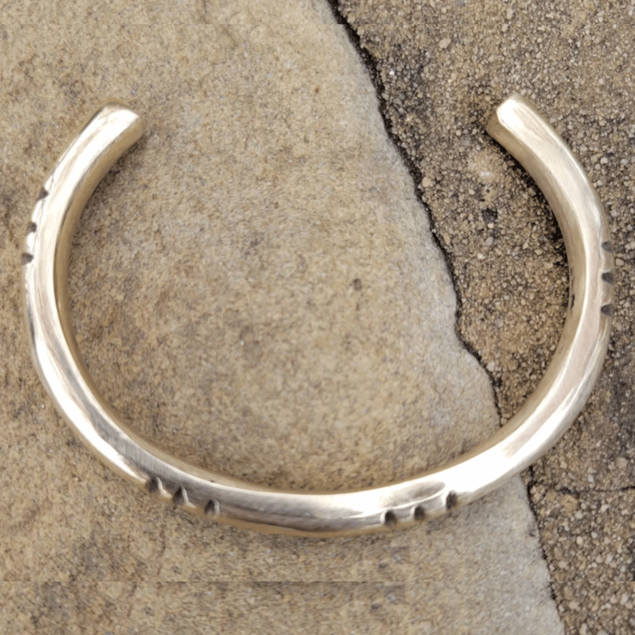 A polished sterling silver cuff bracelet with hand-stamped geometric details, displayed on a textured stone surface. Shop Kamawe