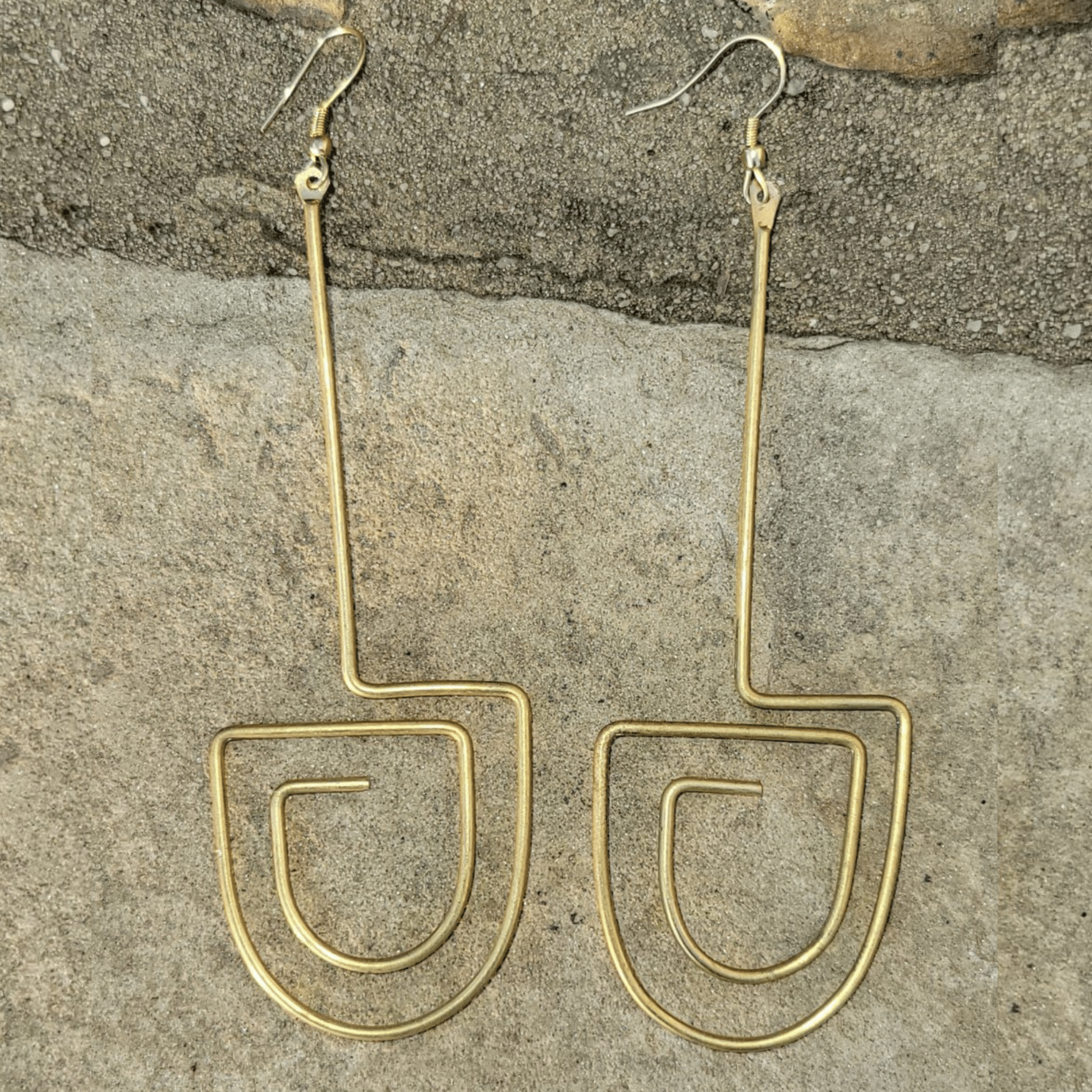 A pair of gold-toned wire dangle earrings with a minimalist, open geometric design, displayed on a textured gray stone surface. Shop Kamawe